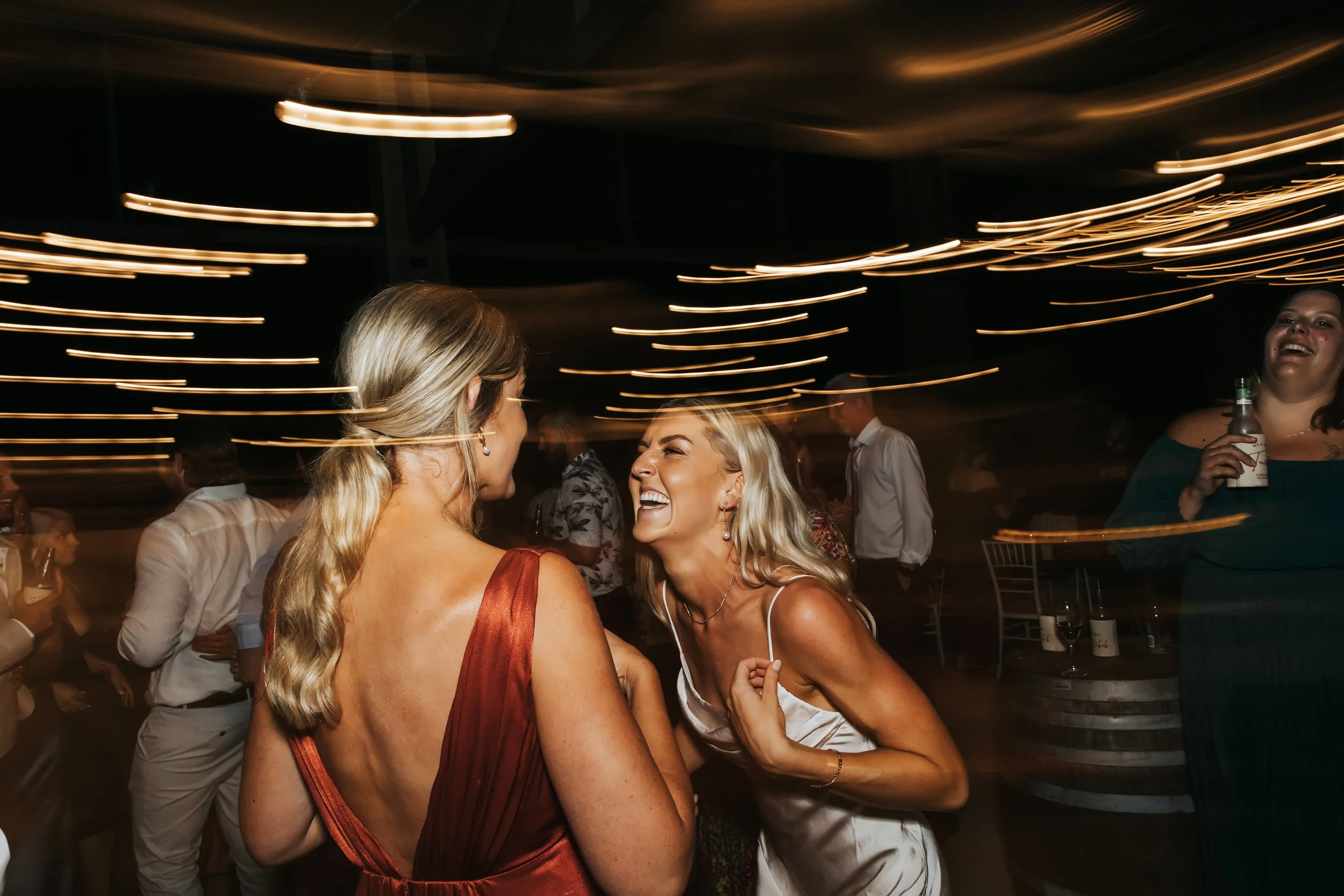 Two women are dancing and laughing at a party or celebration, with other guests in the background. The ceiling has streaks of warm lighting, creating a lively atmosphere.