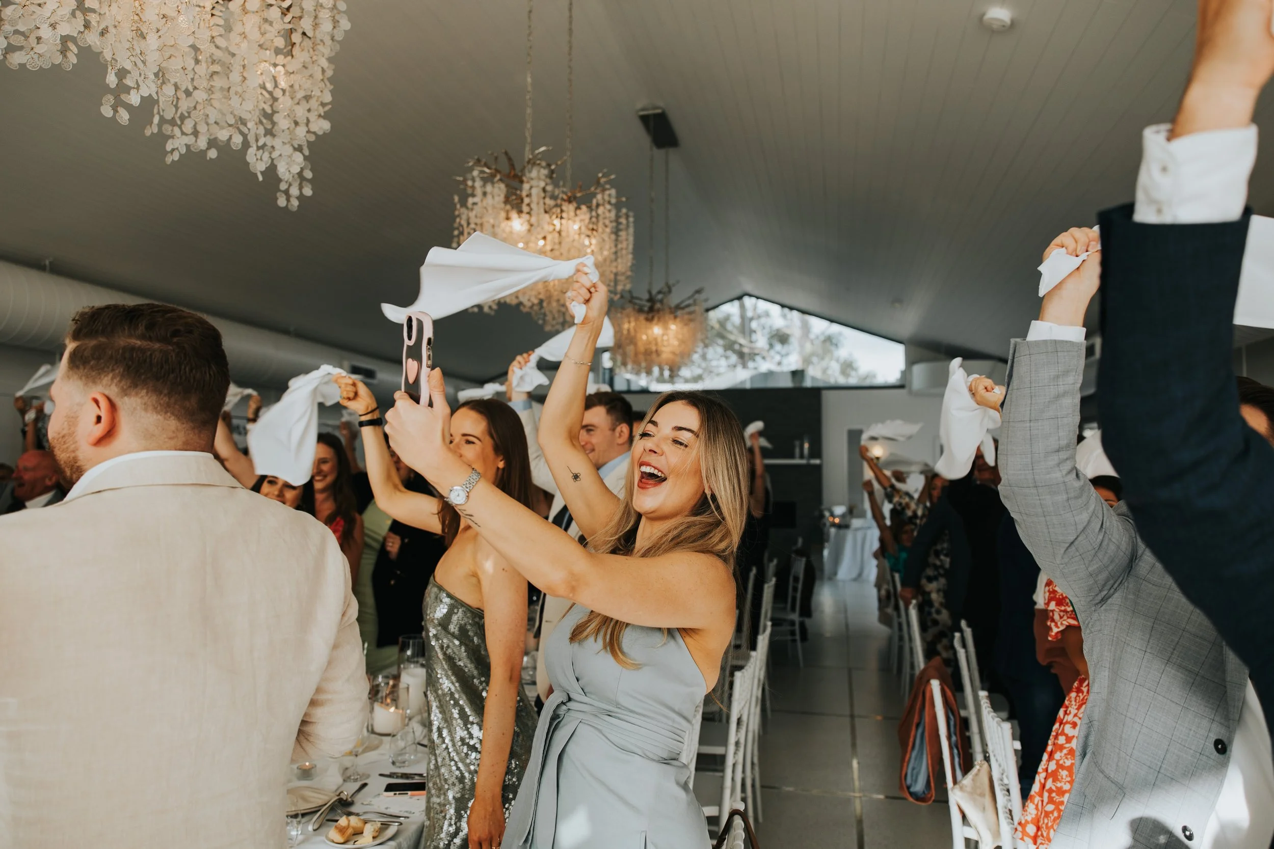 People celebrating, some waving white napkins, in a decorated indoor space with chandeliers.