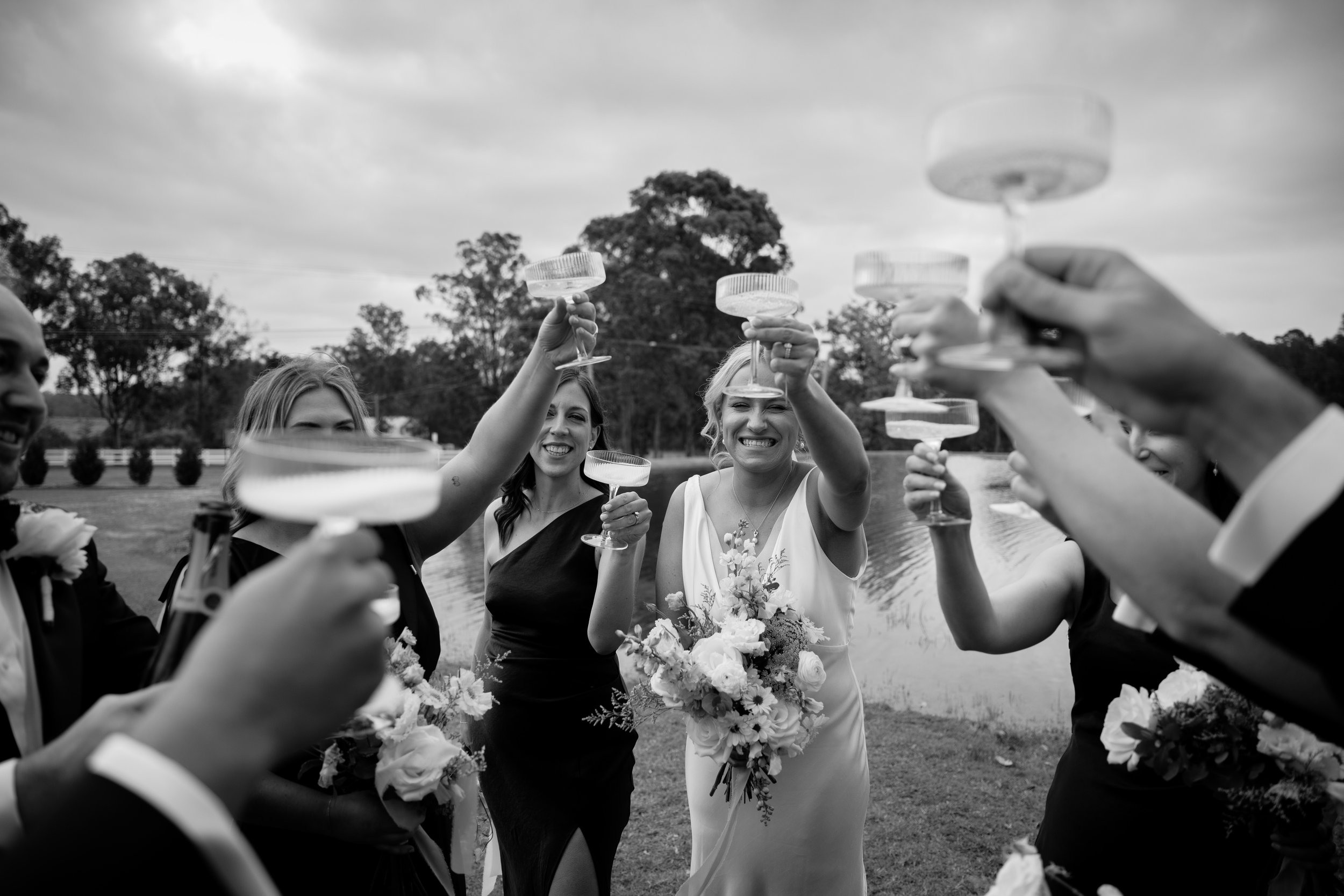 A group of people, including a woman in a white dress holding a bouquet, are raising glasses in a toast outdoors, with trees and a river in the background.