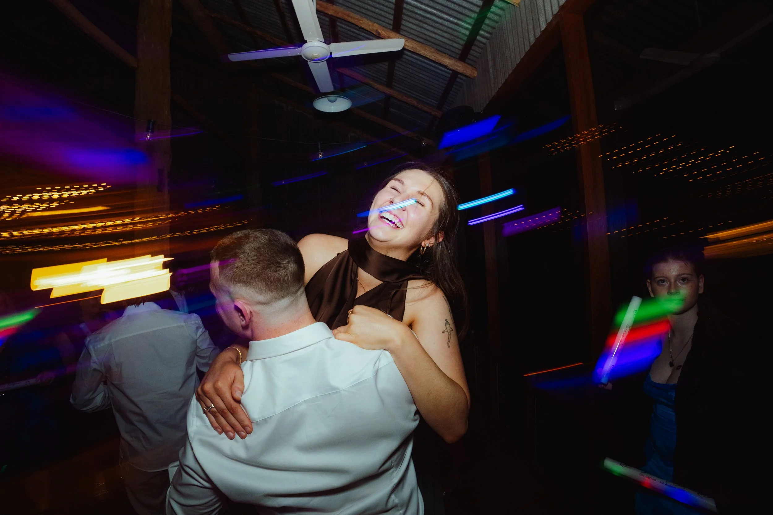 People dancing and celebrating at a party with colorful lights, including a woman smiling and hugging a man, in a rustic venue with a ceiling fan and wooden beams.