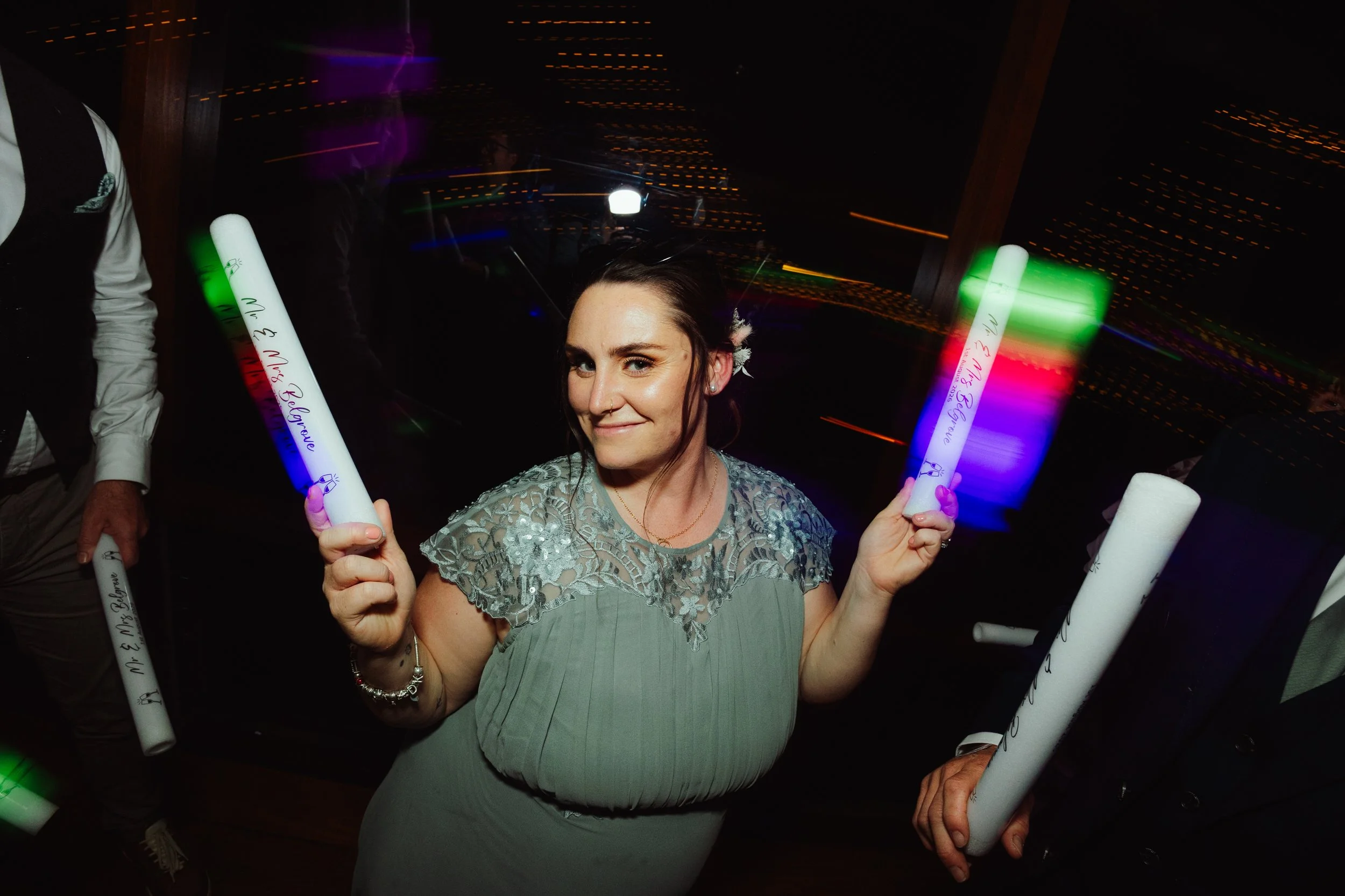 A woman smiling at a celebration, holding light-up glow sticks in a dim, decorated venue.