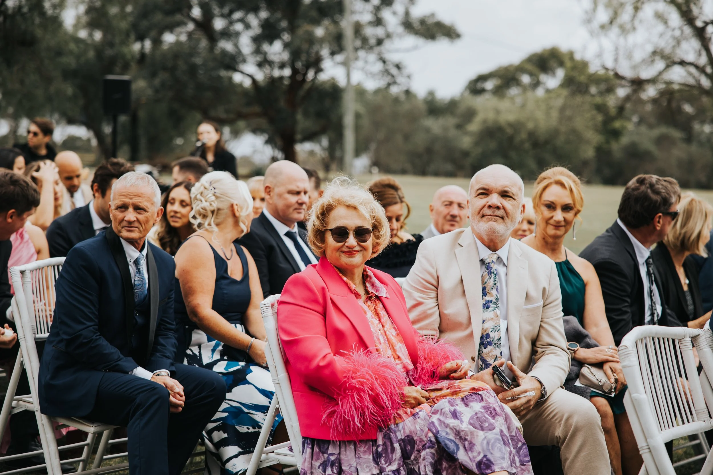 Group of people sitting outdoors at a wedding or formal event, with trees and a field in the background.