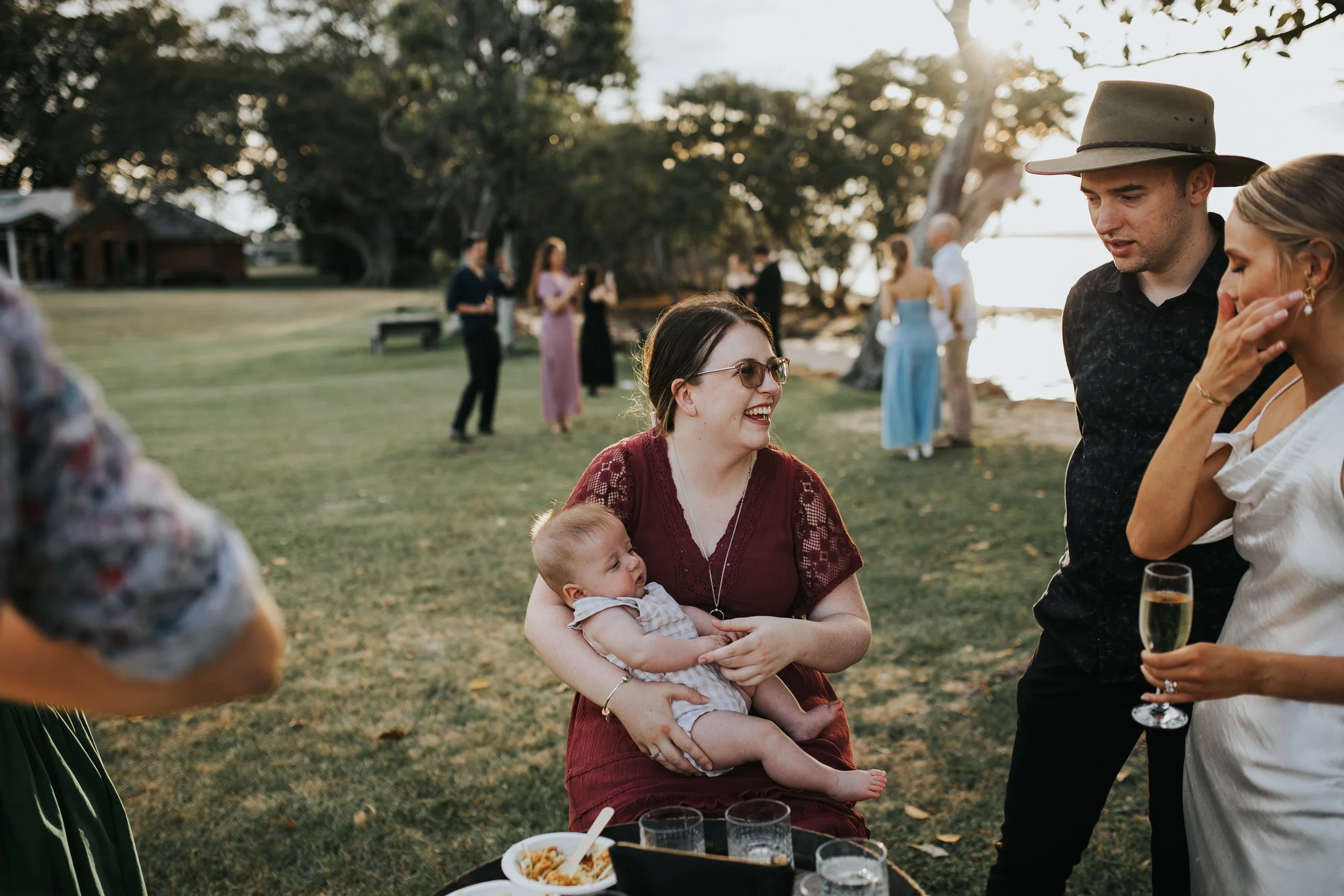 A woman wearing sunglasses holds a baby while smiling, surrounded by people at an outdoor gathering near a body of water during sunset, with trees and a grassy area in the background.