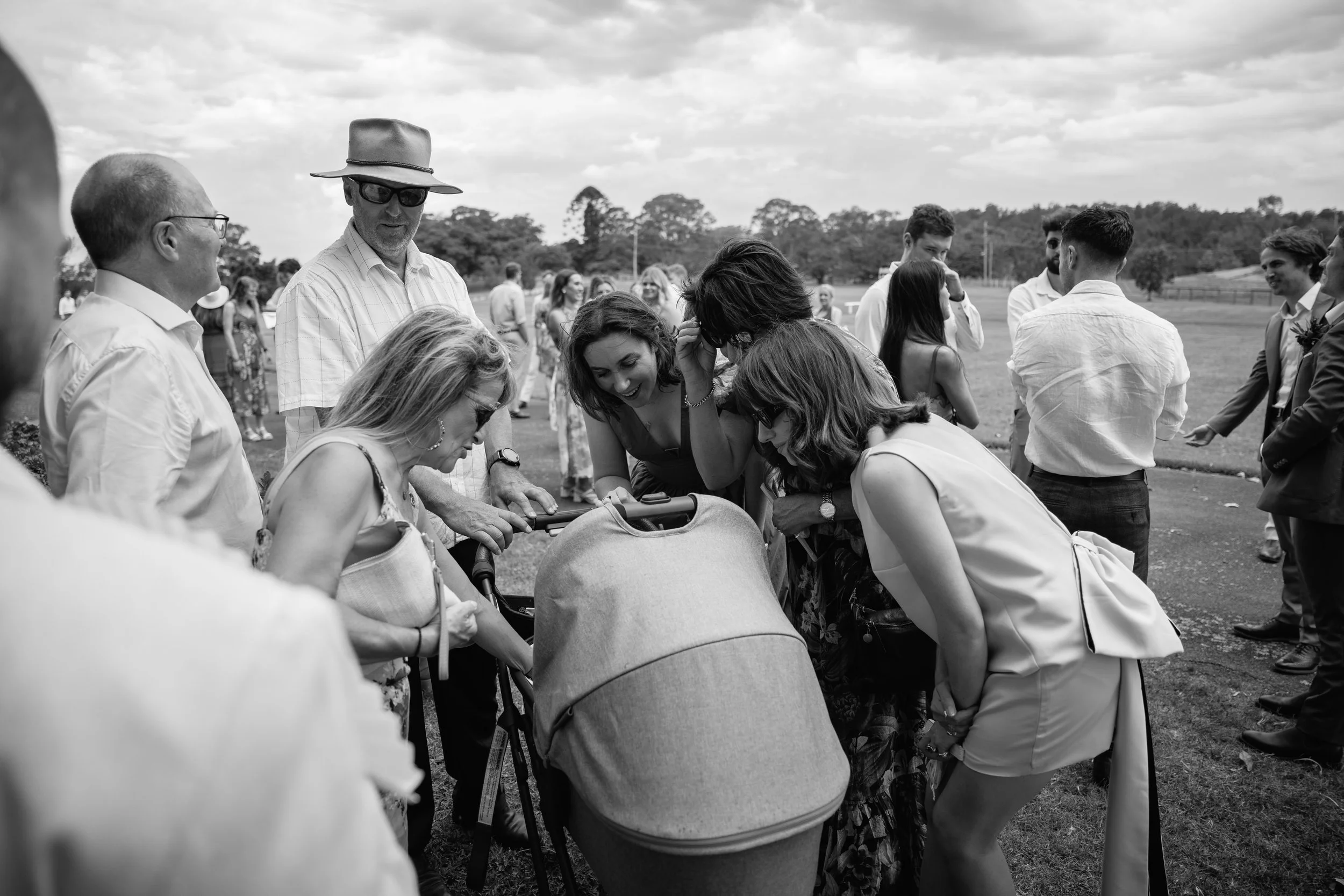 People gathered outdoors, looking into a stroller, with some smiling and engaging, on a cloudy day in a park or open field.