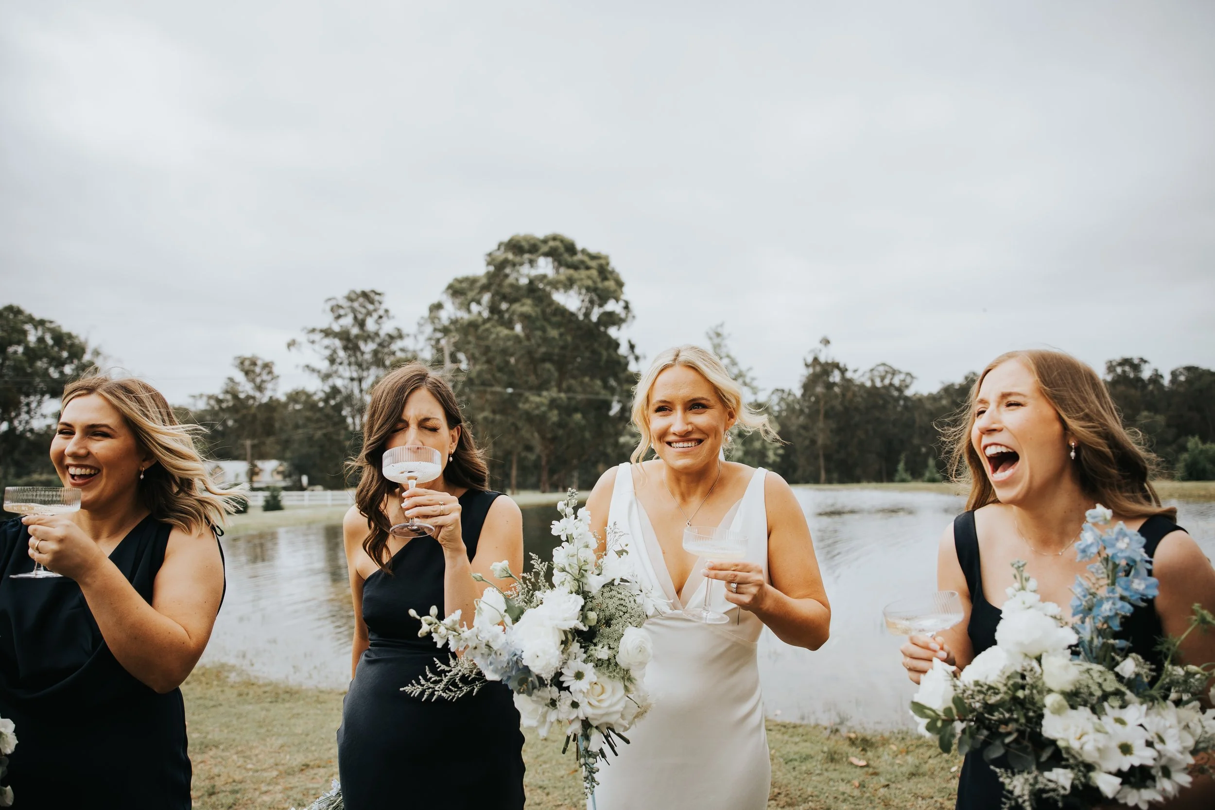 A group of women in formal dresses celebrating outdoors near a body of water, holding glasses and flowers, with trees and cloudy sky in the background.