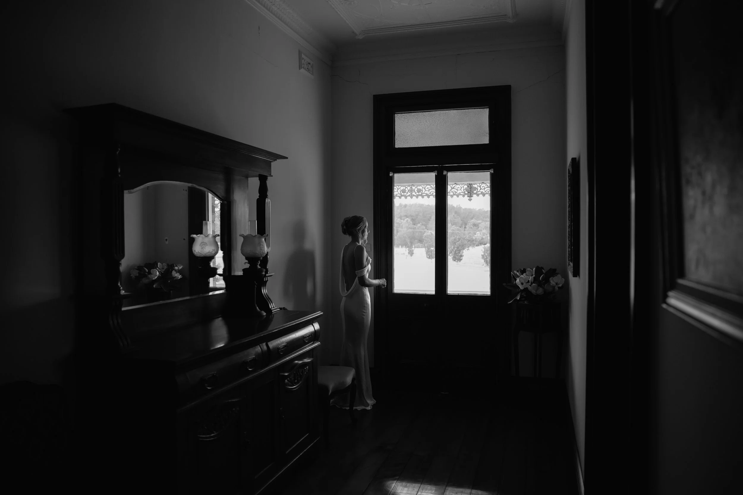 A woman in a dress stands near a large window with a view of trees outside in a dimly lit room with antique furniture and decorative items.