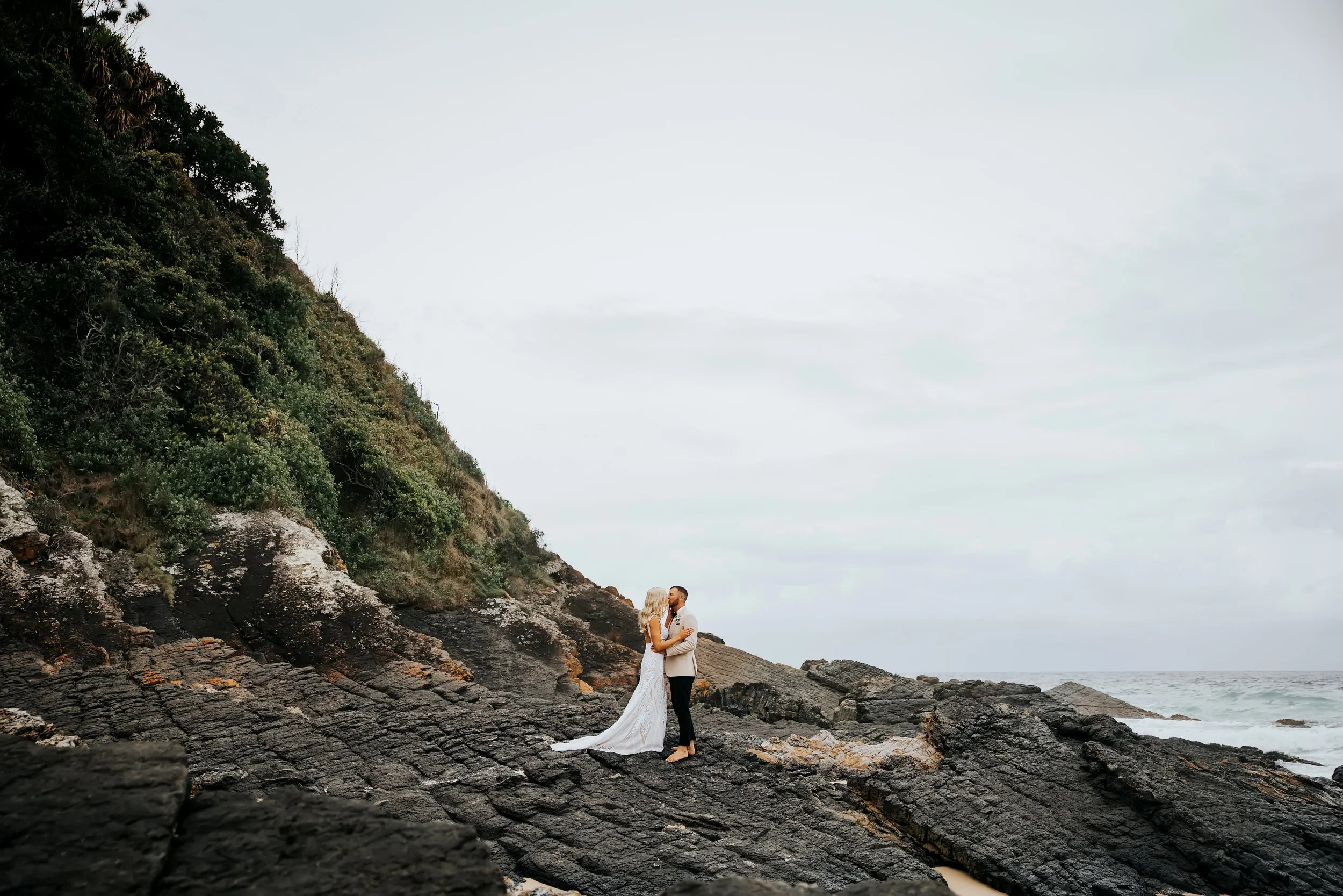 A couple in wedding attire embracing on rocky shoreline with ocean and cliffside greenery in the background.
