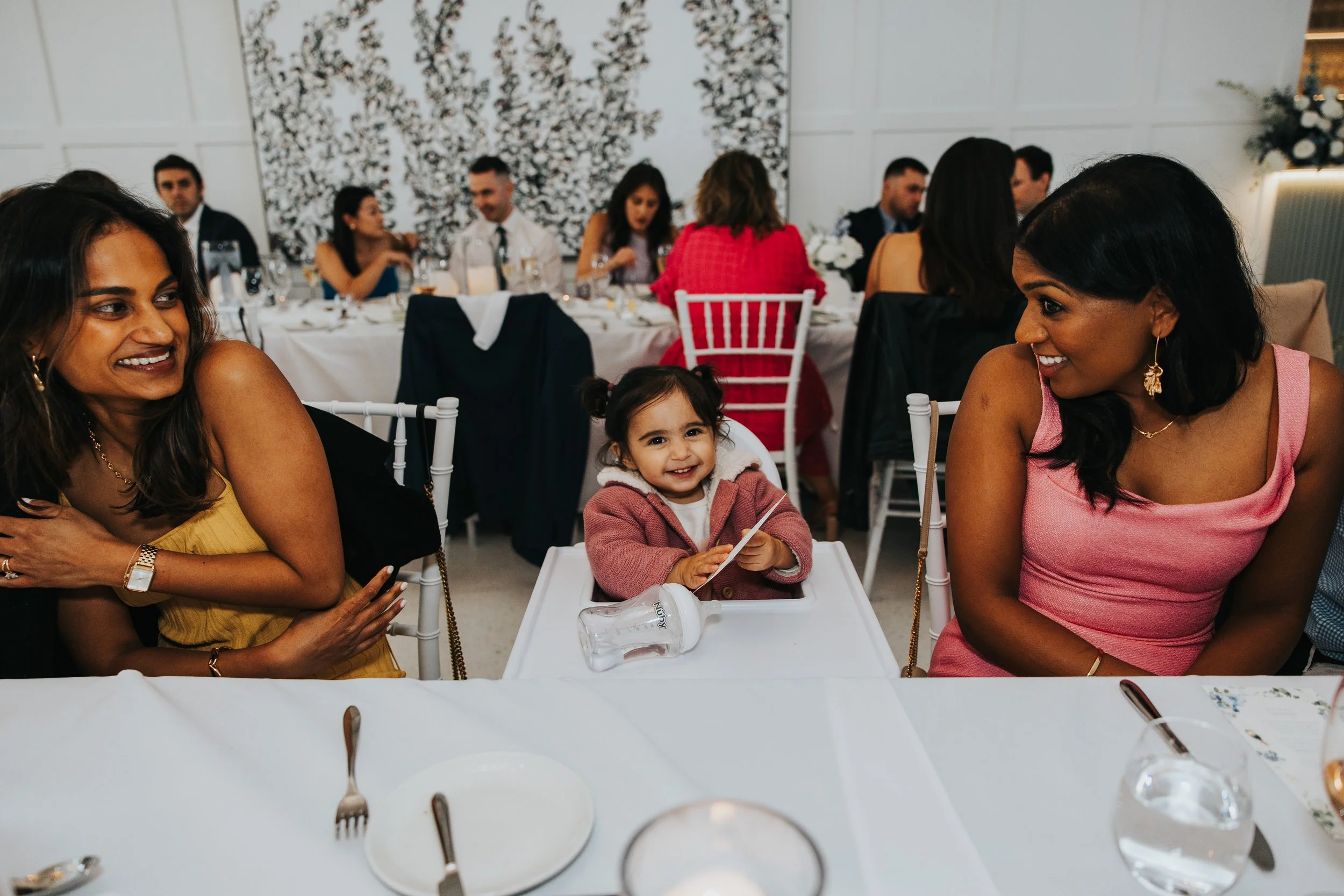 People gathered at a formal event, sitting at a table with dinnerware, in a decorated room with white walls and floral art. Two women and a young girl are in the foreground, smiling and talking, with others in the background engaged in conversation.