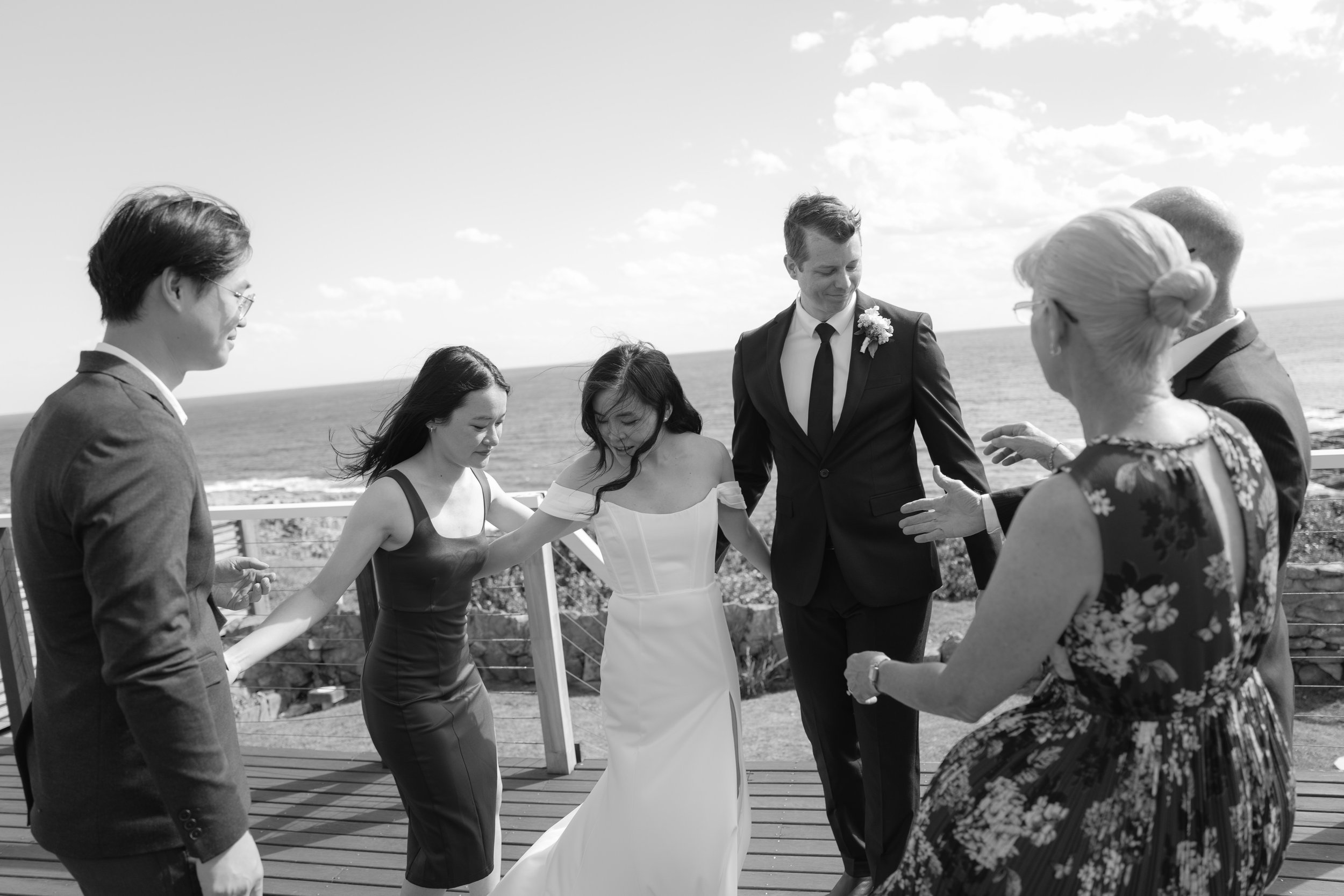 A black and white photo of a wedding ceremony outdoors by the ocean, with a bride, groom, and several guests holding hands in a circle.