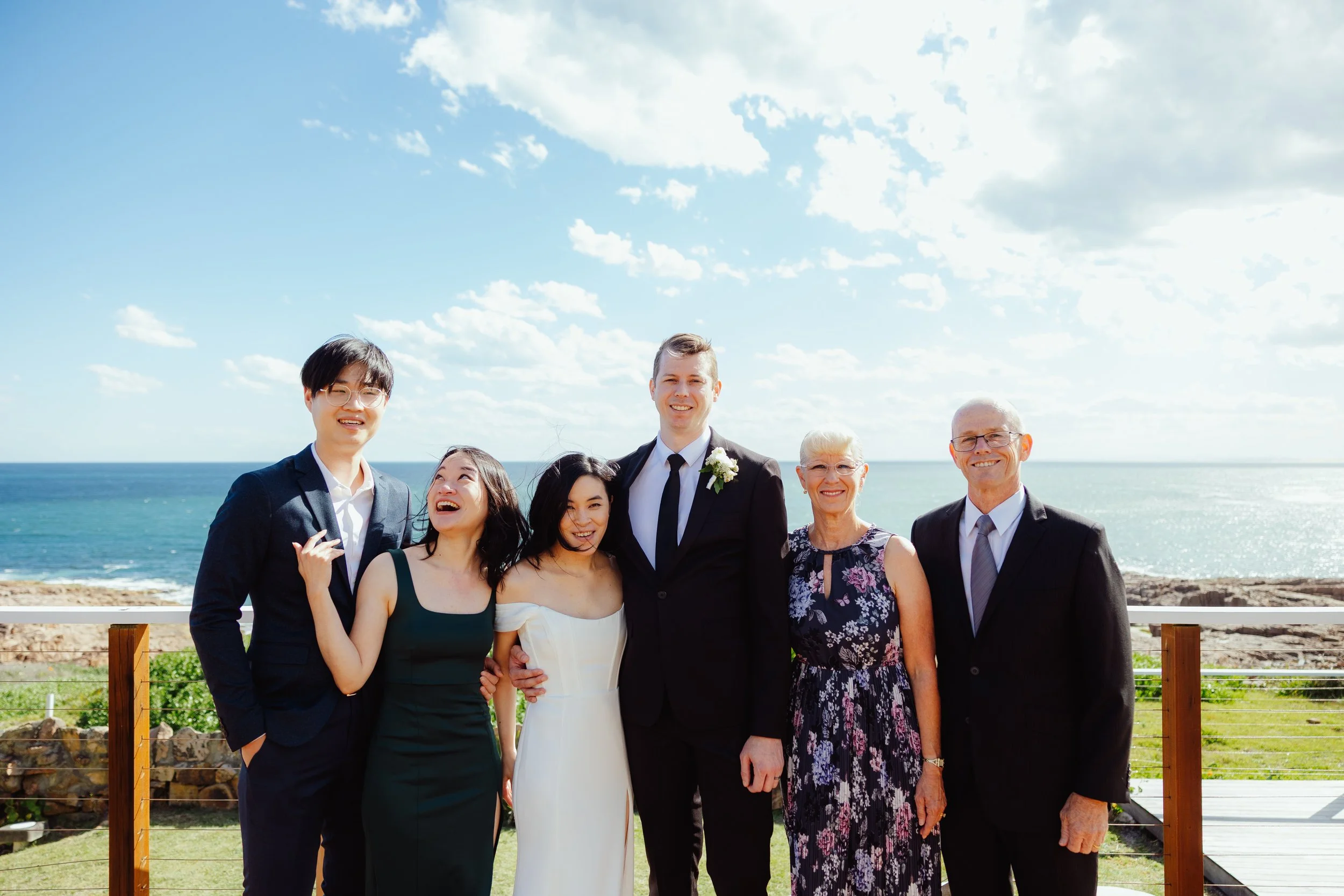 Group of six people standing outdoors near the ocean under a partly cloudy sky, dressed in formal attire, smiling for a photo.