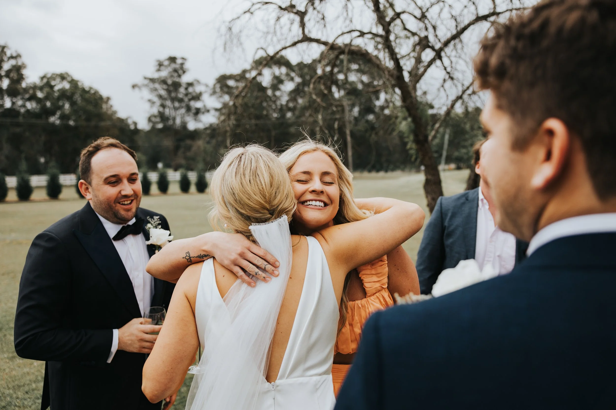 Group of people at an outdoor wedding, hugging and smiling, with trees and a fence in the background.