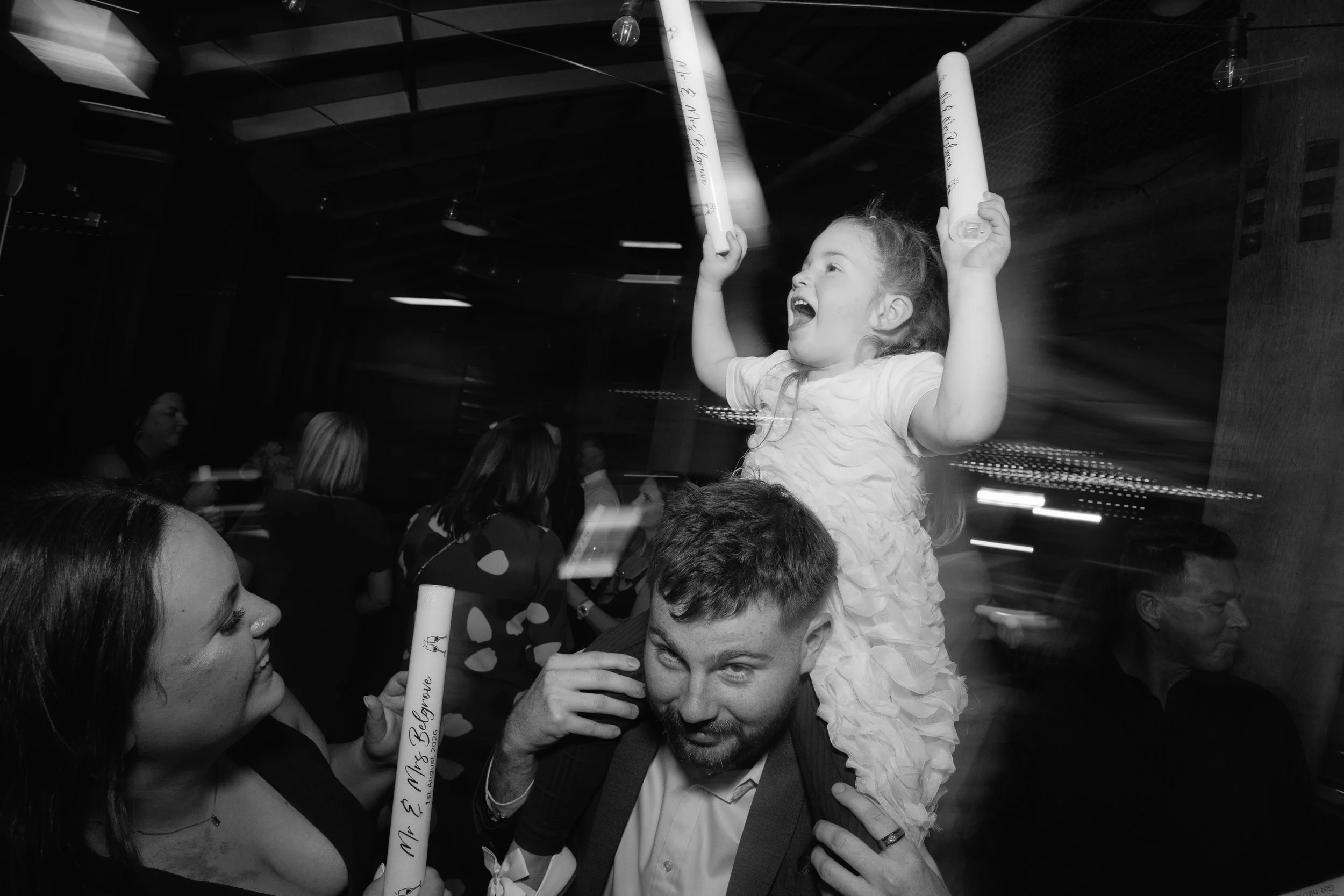 A joyful young girl with curly hair cheers and holds a bat while sitting on a man's shoulders at a celebration event. Other people are visible in the background.