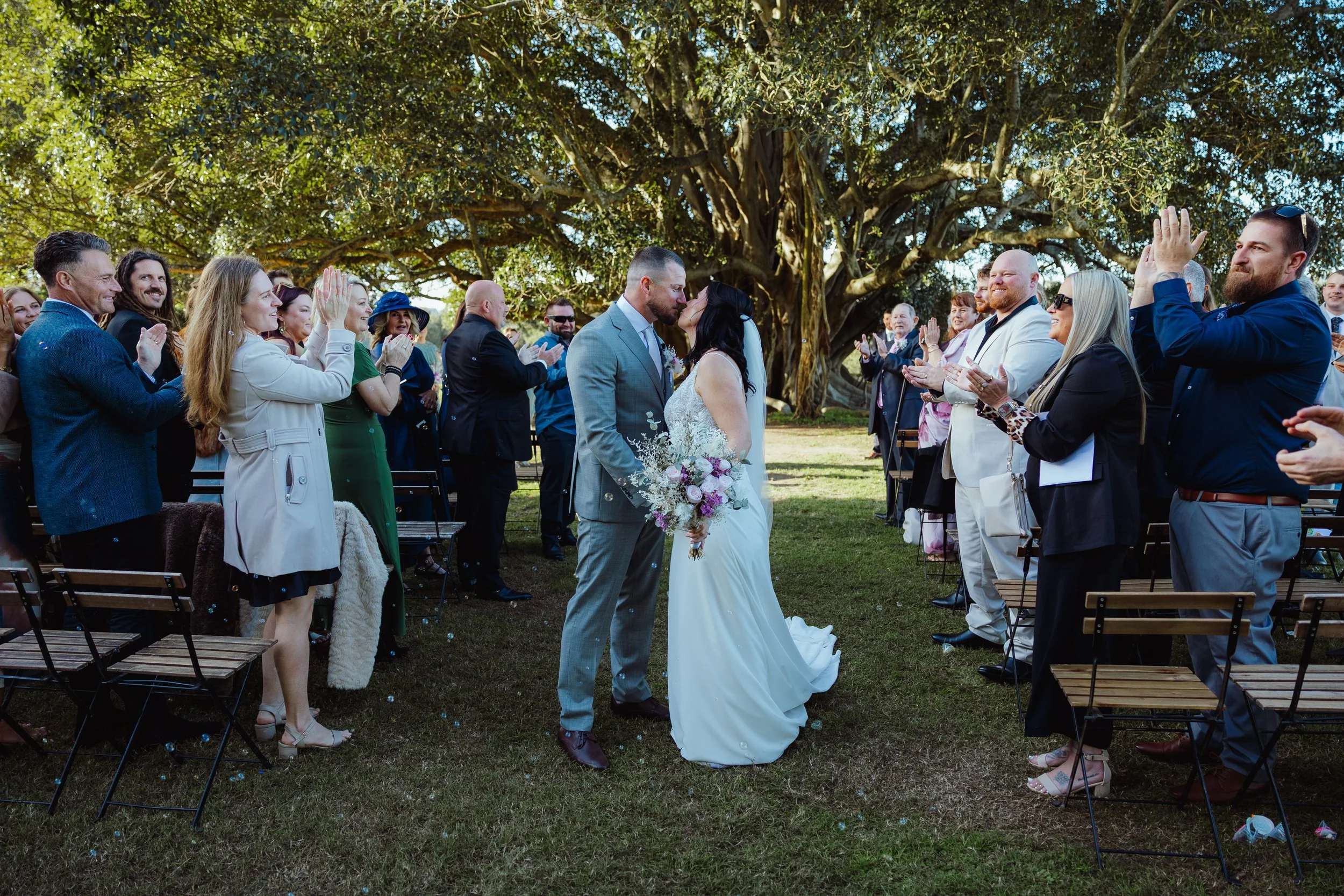 A bride and groom kiss during their outdoor wedding ceremony under a large tree, with guests applauding around them.