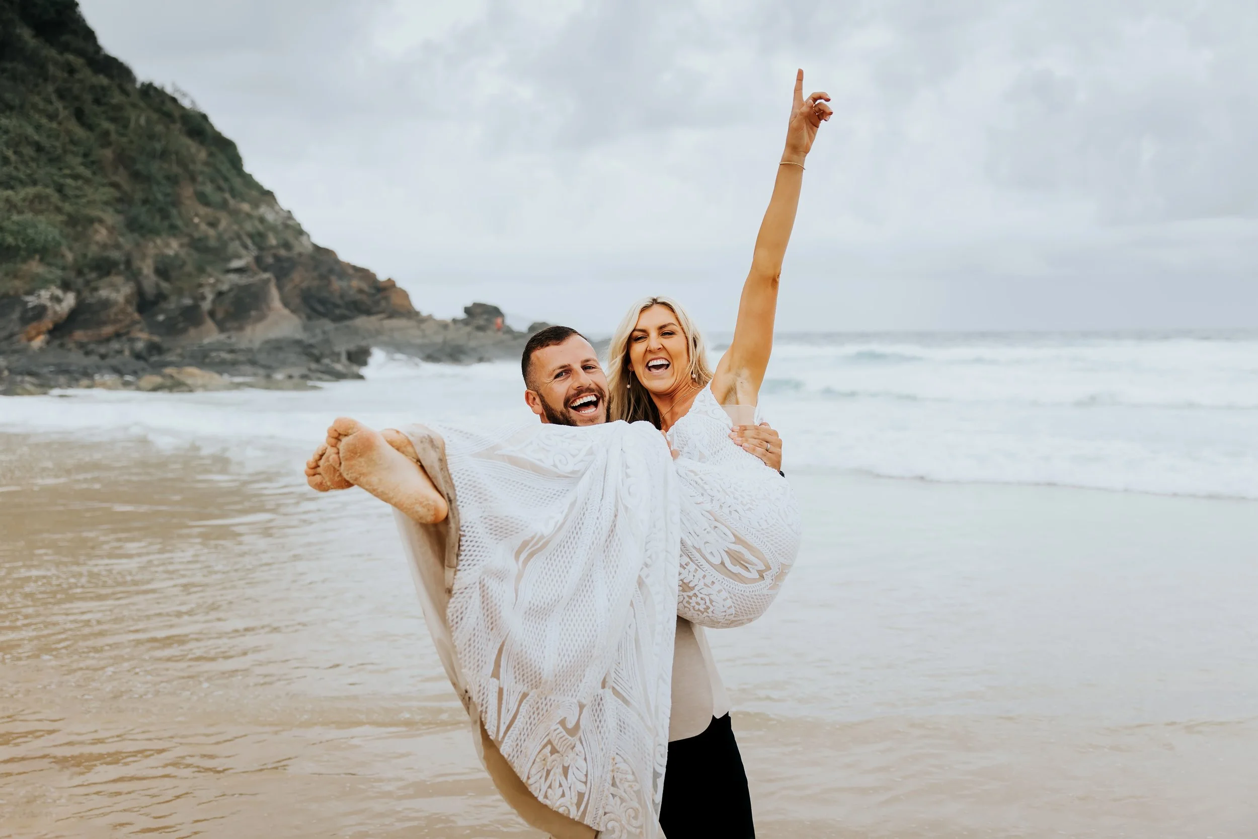 A happy couple on the beach, with the woman raising one arm and smiling, as the man holds her, near the shoreline with rocks and green hills in the background.