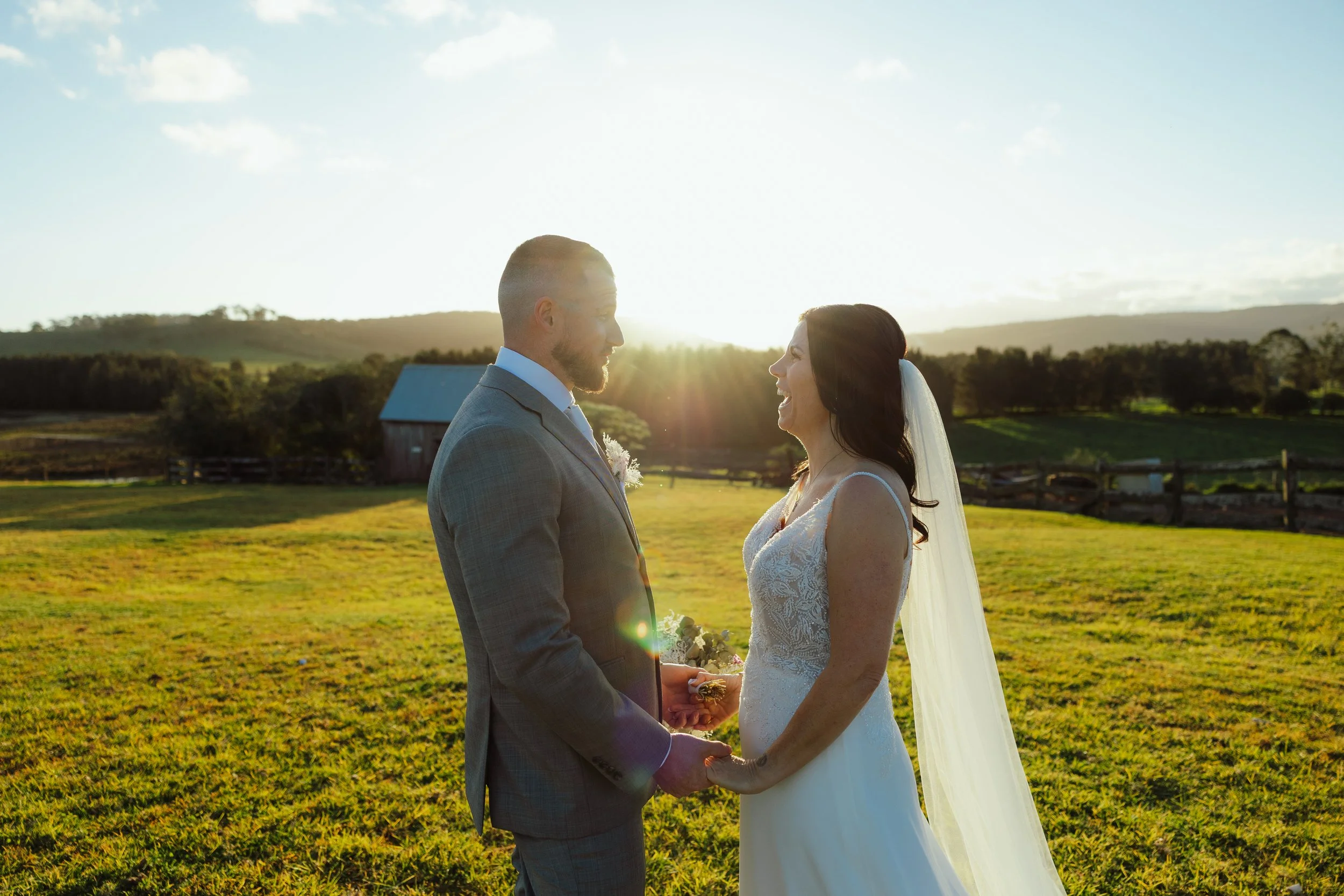 A bride and groom holding hands and facing each other outdoors during sunset, with a farm and trees in the background.