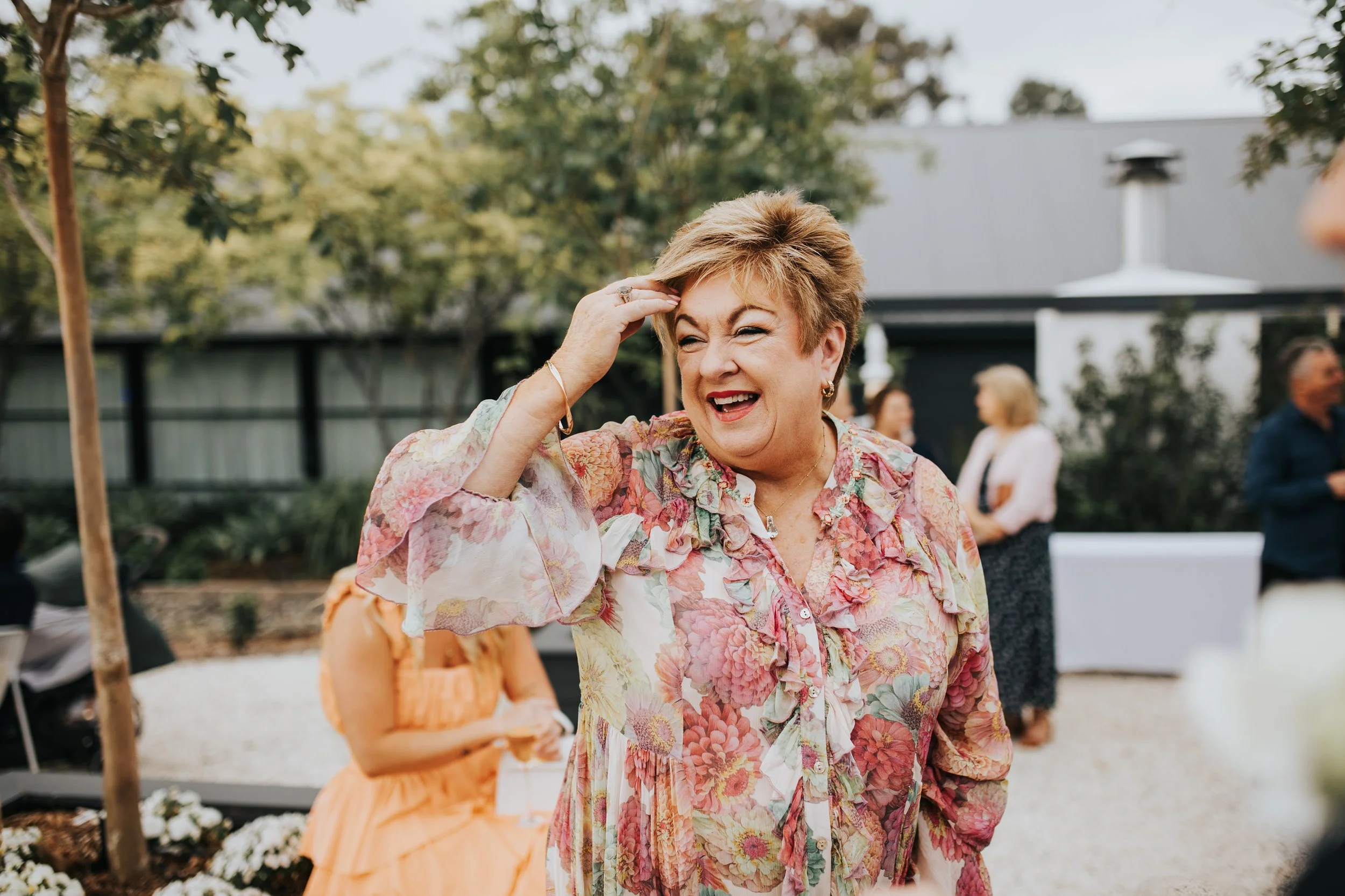 A smiling woman with short, light brown hair, wearing a floral blouse, is holding her hand to her head outdoors during daytime. In the background, there are other people and a building with a chimney, trees, and decorative plants.