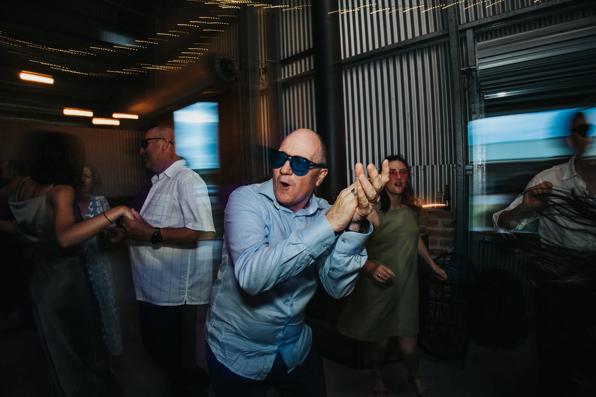 A group of people dancing and socializing at an indoor event with string lights overhead, featuring a man with sunglasses in the foreground and others of different ages engaged in conversation and dancing.