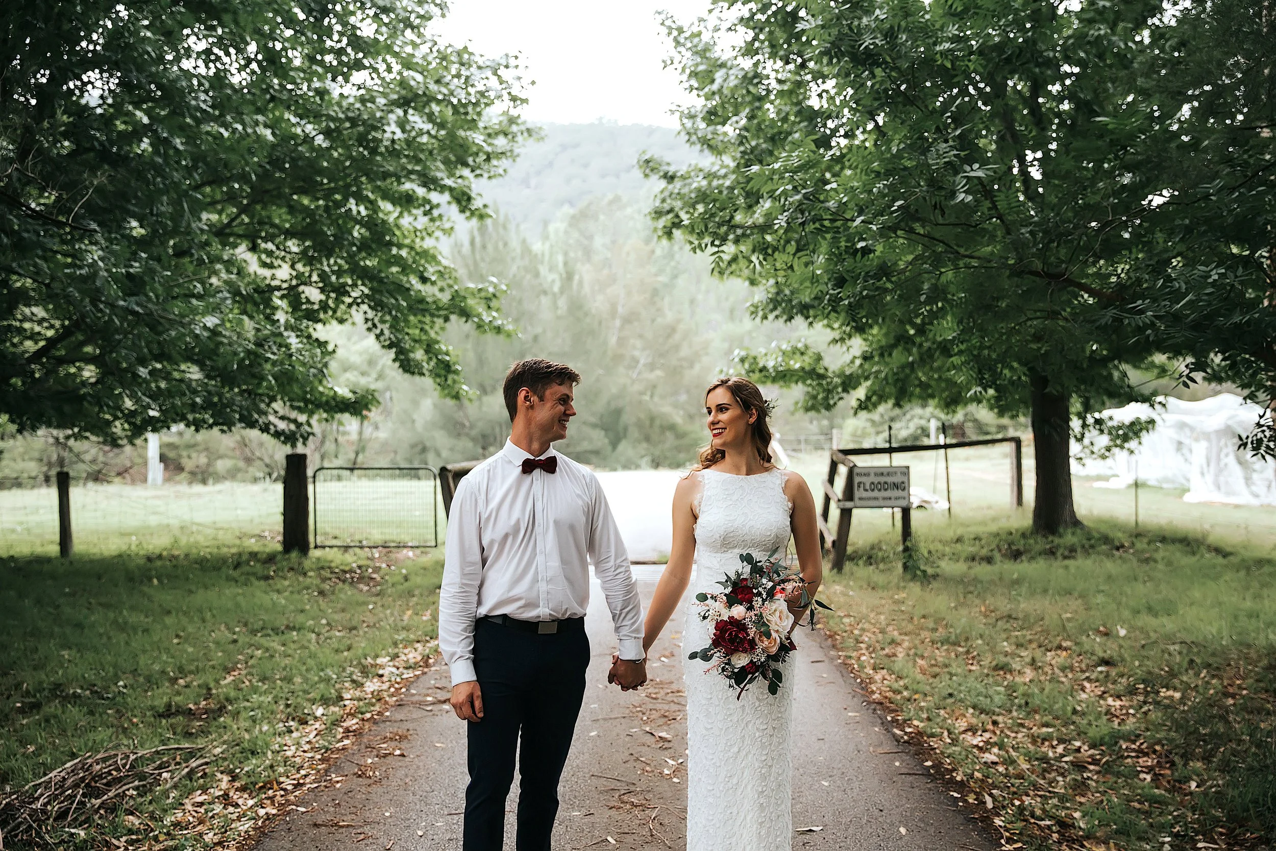 A newlywed couple walking hand in hand outdoors in a lush green park, holding a bouquet of flowers, with trees on either side and a gate in the background.