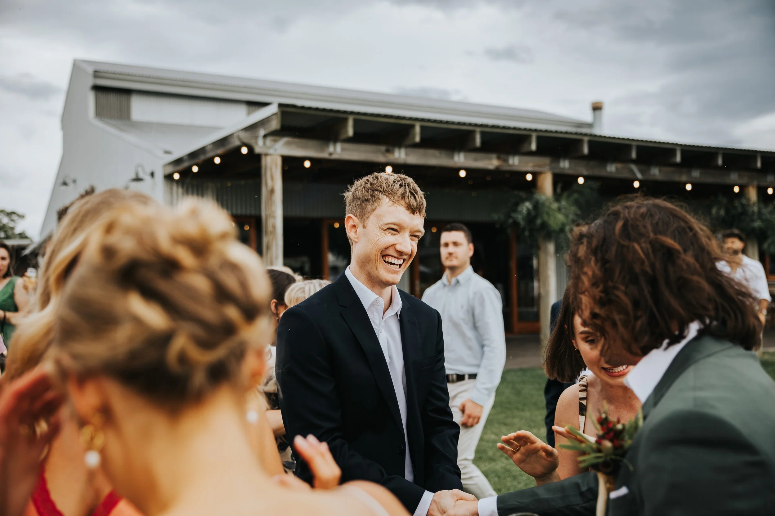 A group of people, including a smiling man in a suit, gathered outdoors at what appears to be a celebration or wedding, with a rustic building in the background under a cloudy sky.