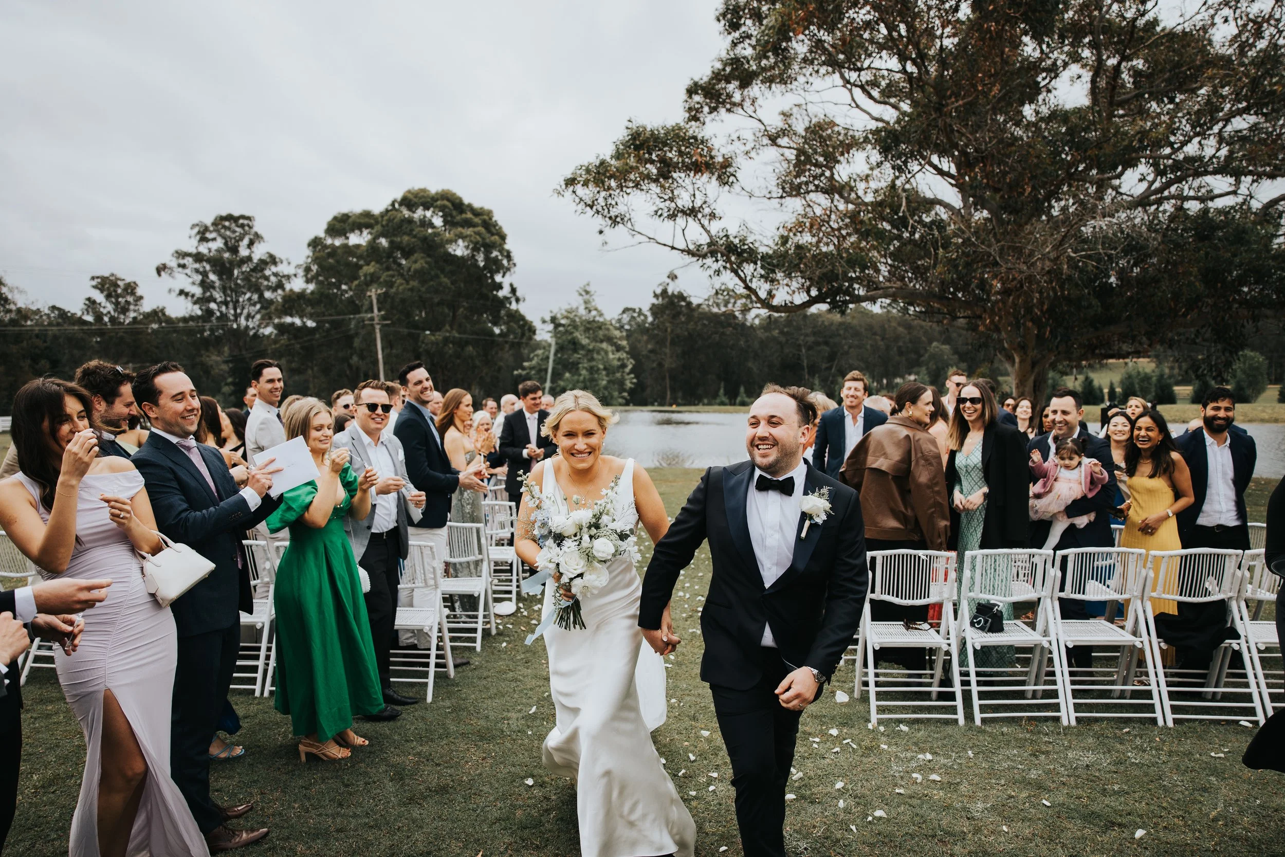 A bride and groom holding hands and walking down the aisle at an outdoor wedding ceremony, surrounded by smiling guests.