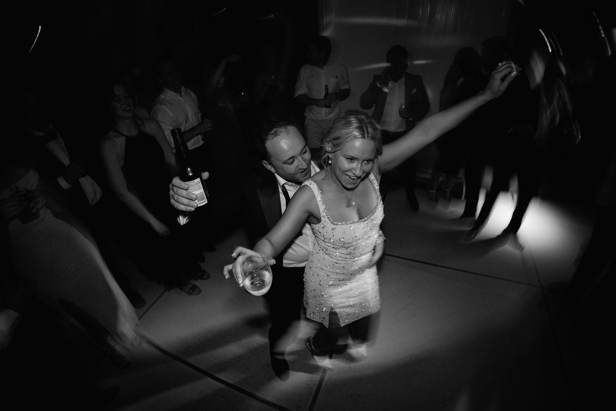 Couple dancing closely at a party with people celebrating around in a dimly lit room.