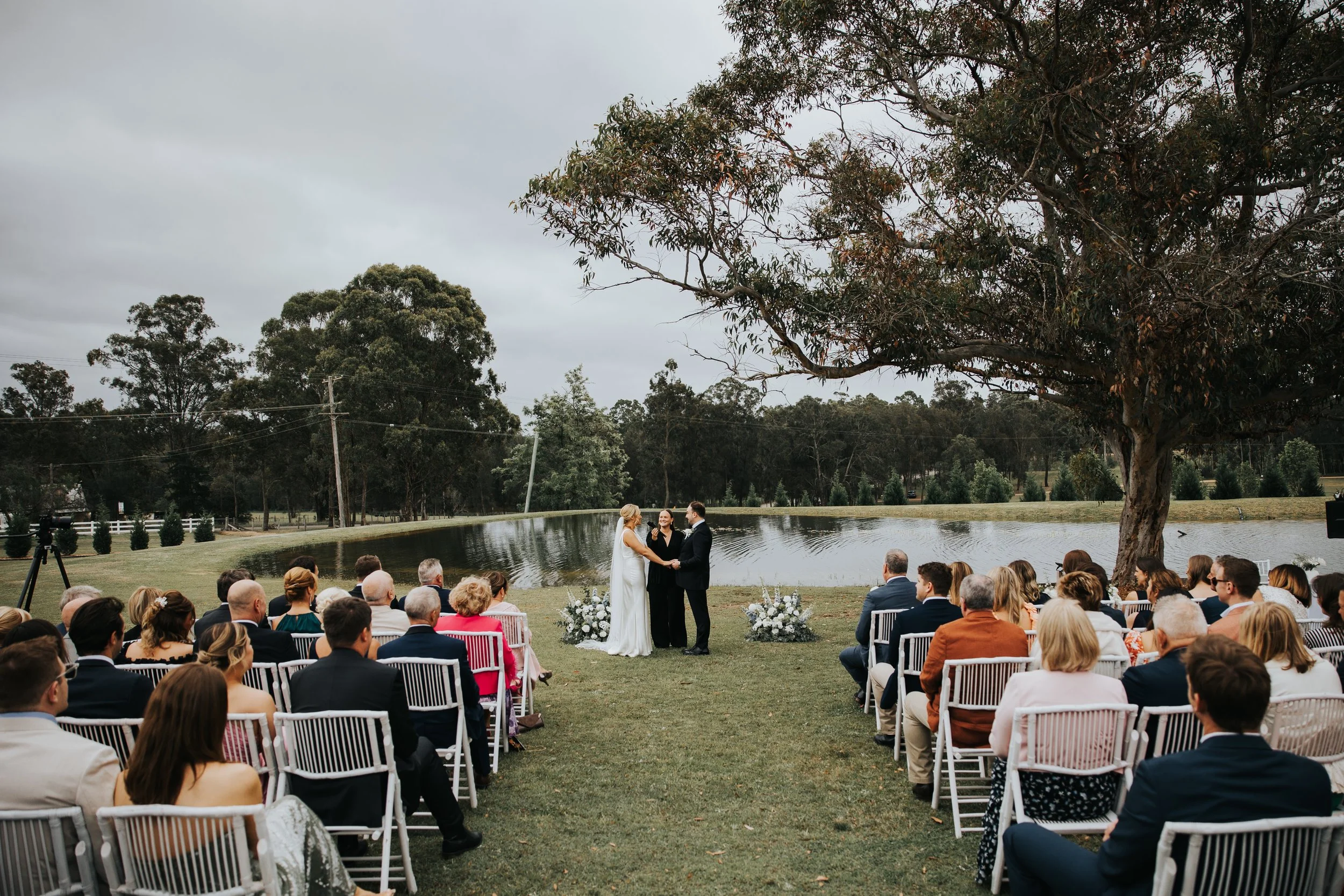 An outdoor wedding ceremony by a pond with guests seated on white chairs, a bride and groom exchanging vows, and a large tree and overcast sky in the background.