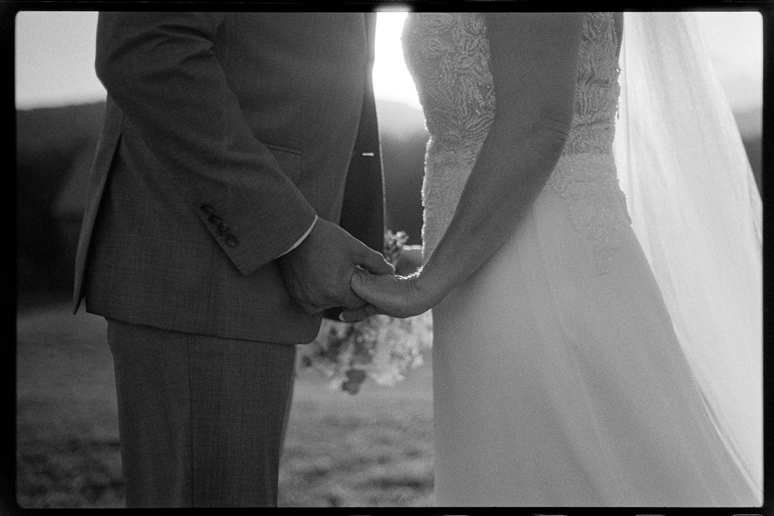 Close-up of a couple holding hands during a wedding ceremony, with the bride wearing a lace wedding dress and the groom in a suit, outdoors in natural lighting.