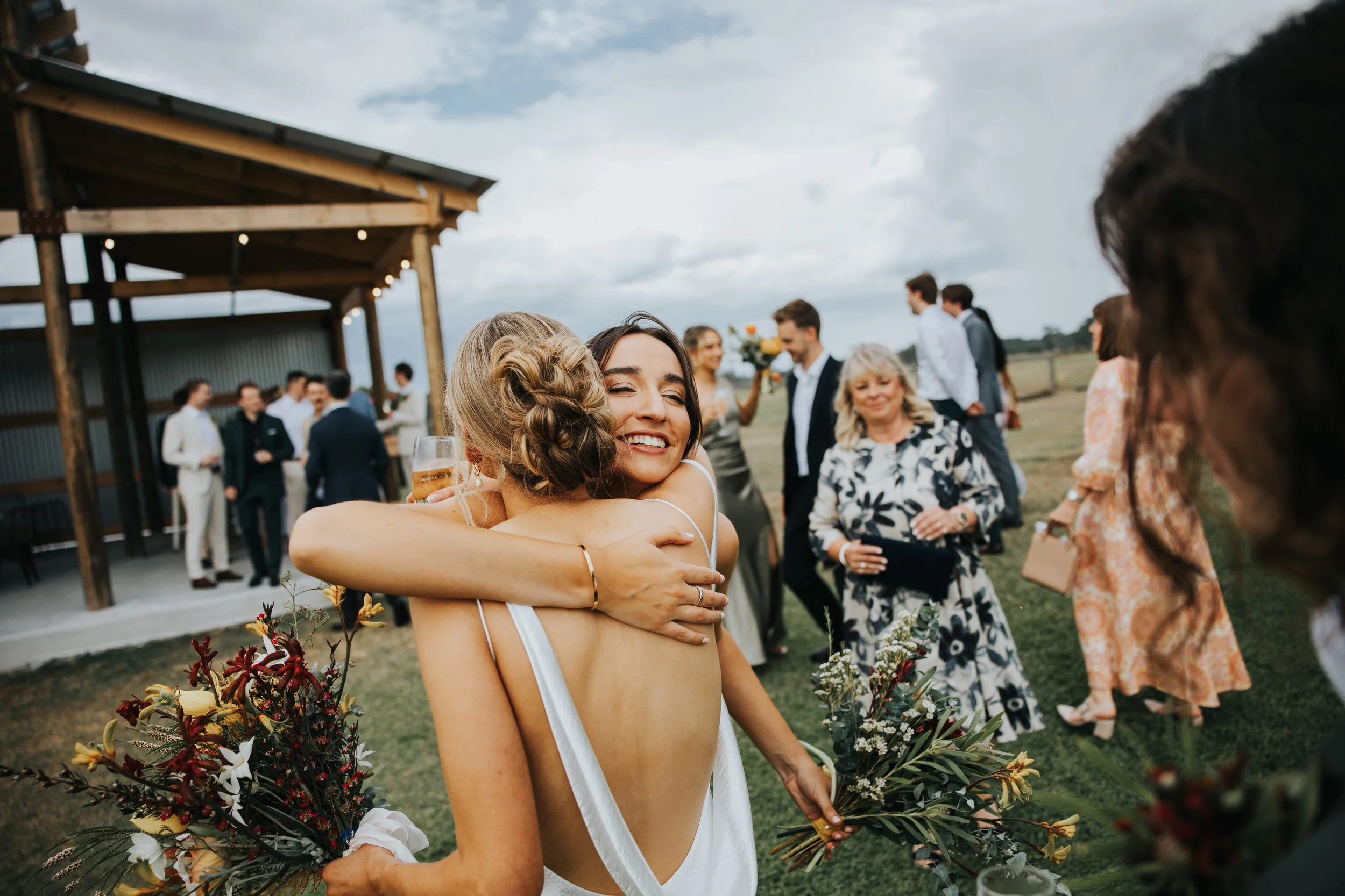 A woman with dark hair and a big smile hugging another woman with blonde hair at an outdoor wedding reception, with other guests and a wooden pavilion in the background.