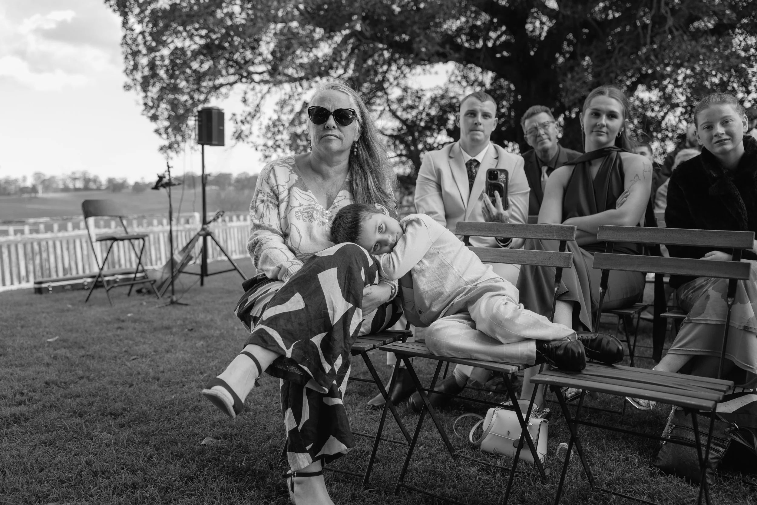A group of people sitting outdoors under a large tree, with a woman in sunglasses and a child resting on her lap in the foreground. Others are looking forward, some with phones, with a fence and open field in the background.