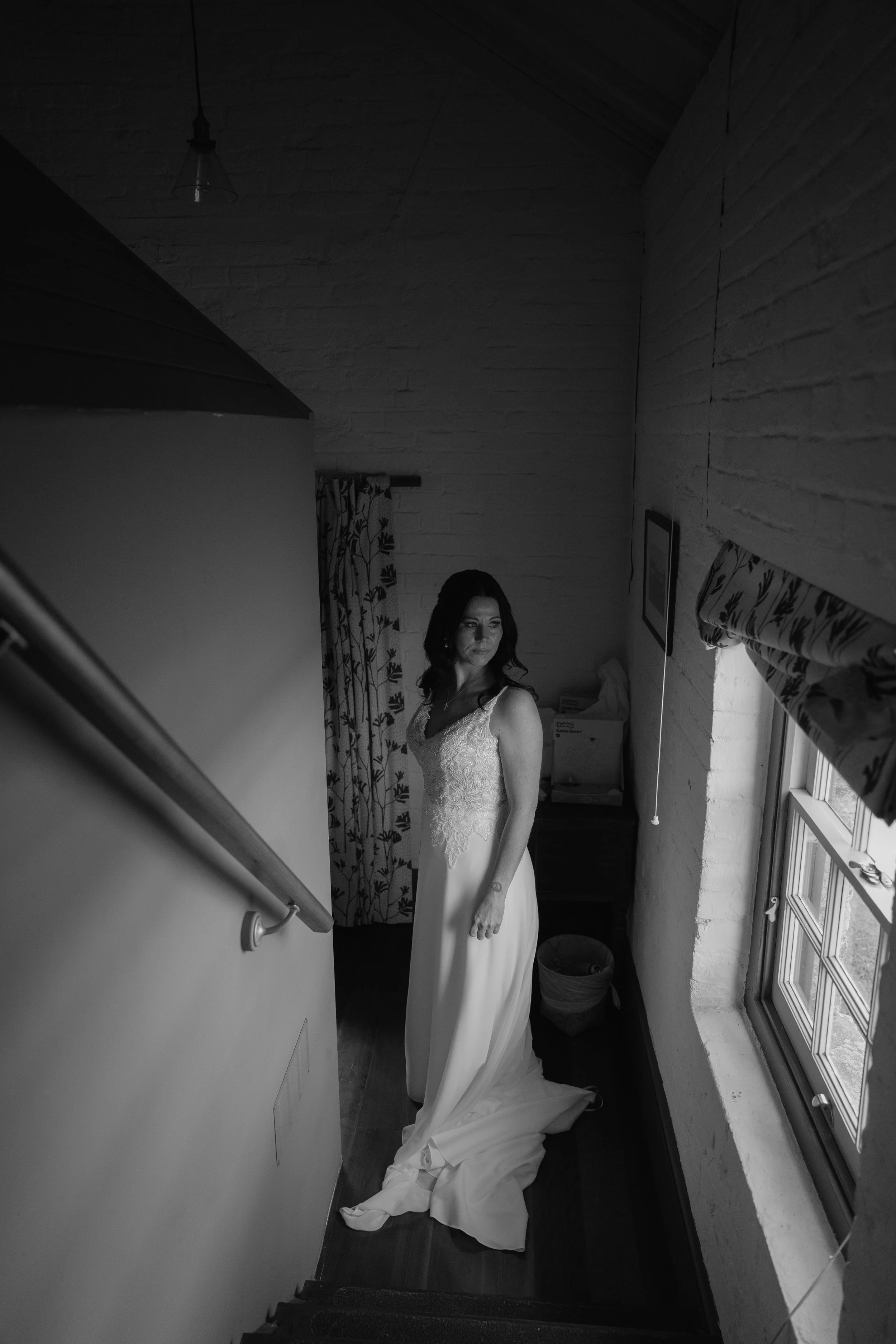 A woman in a wedding dress standing in a room with brick walls, a window with curtains, and a ceiling light. The dress has a lace bodice and a long train that pools on the floor.