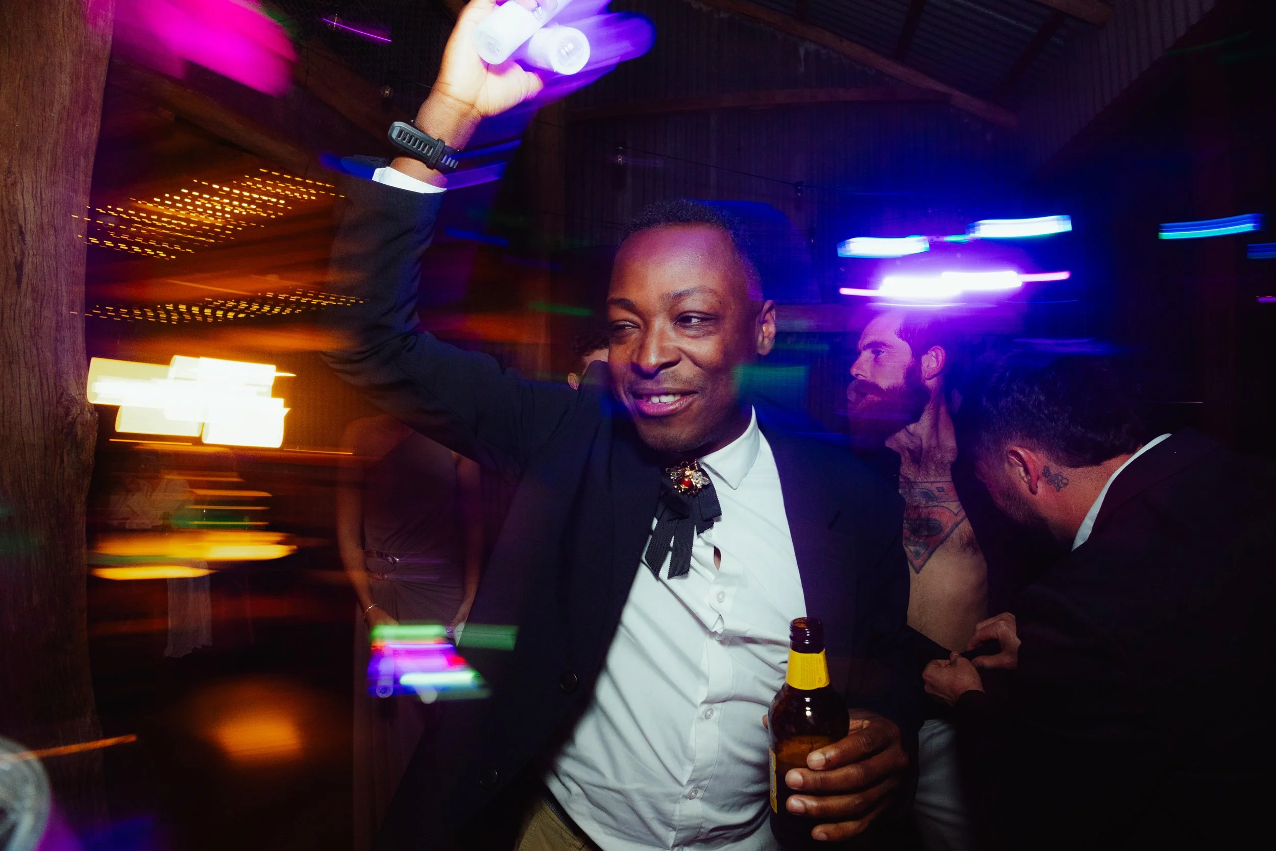Man wearing a tuxedo with a bow tie and a brooch, holding a beer bottle, at a party with colorful lights creating motion blur.