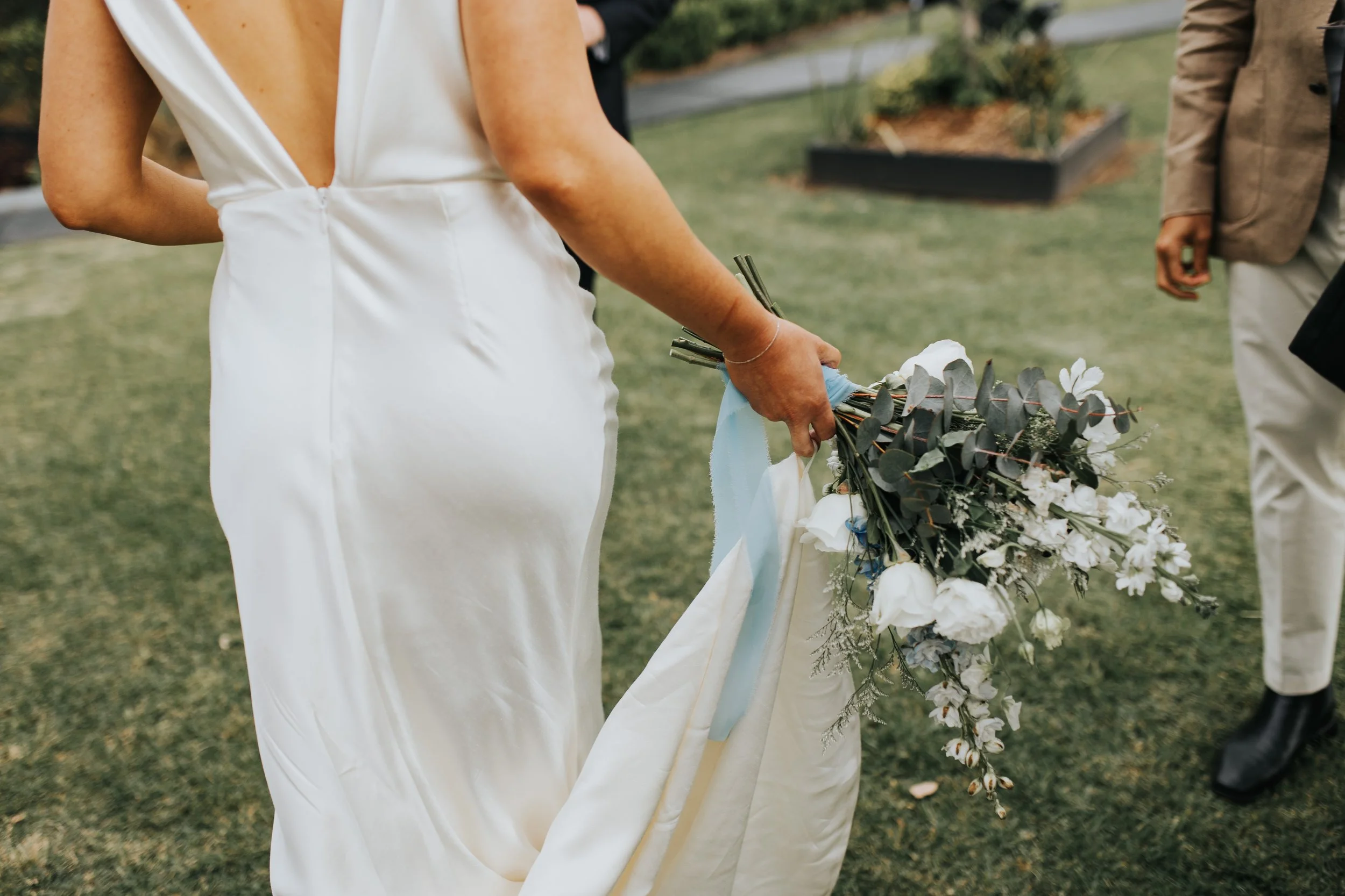 A bride in a white dress holding a wedding bouquet with white flowers and greenery at an outdoor wedding ceremony.
