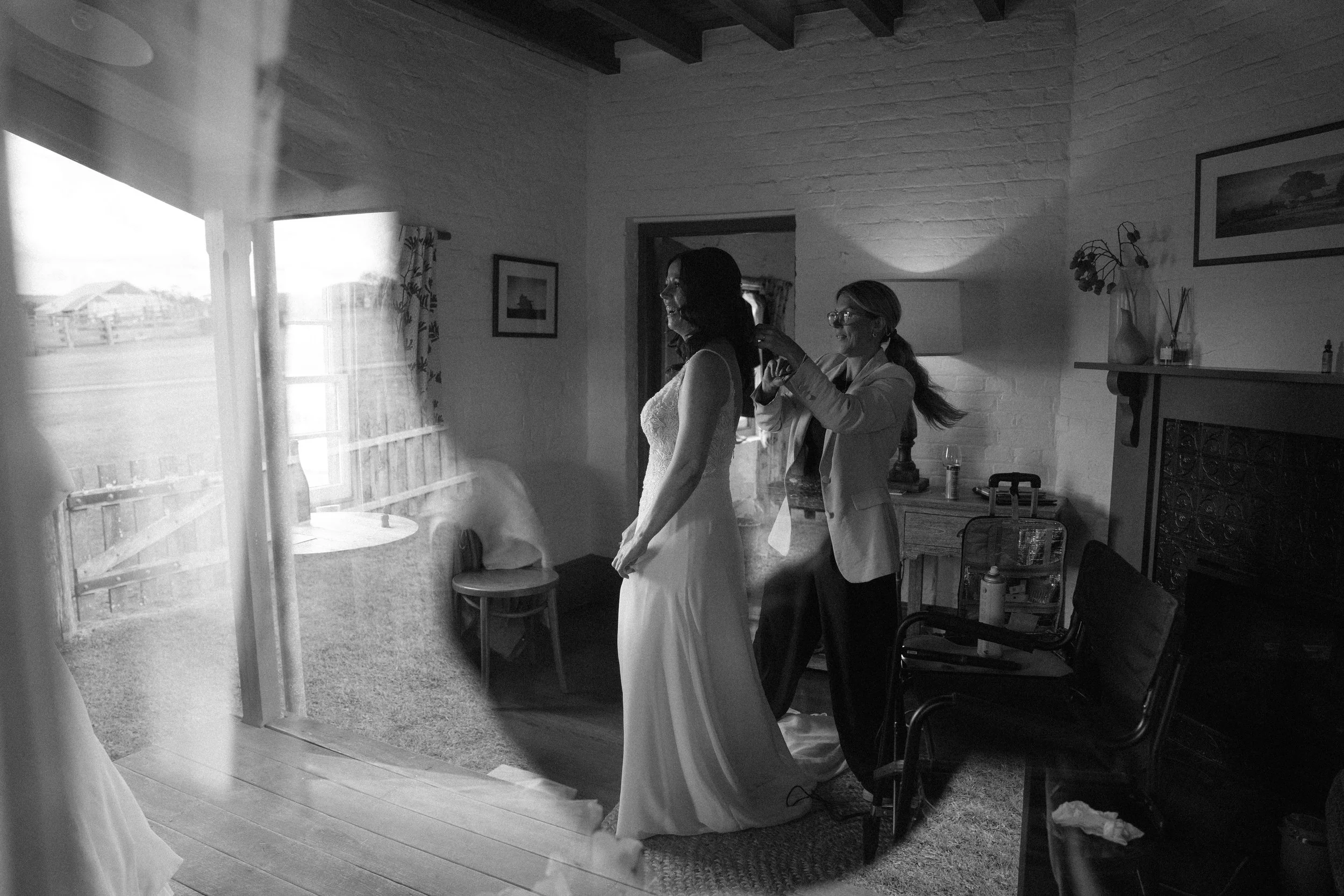 A woman in a wedding dress getting ready with the help of another woman in a cozy, well-lit room with a fireplace, framed pictures on the walls, and large windows showing an outdoor view.