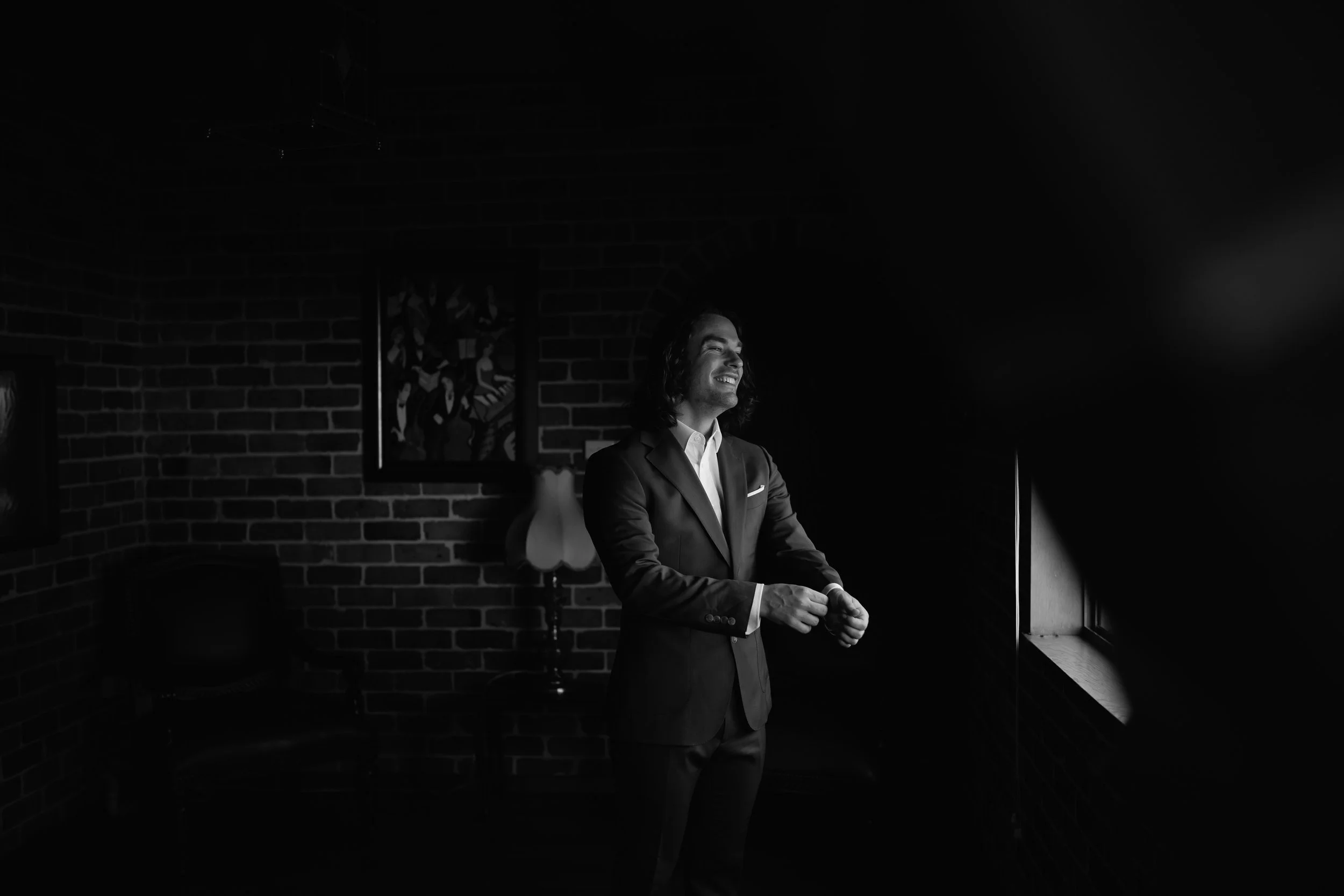 A man in a suit smiling while sitting by a window in a dimly lit room with brick walls.