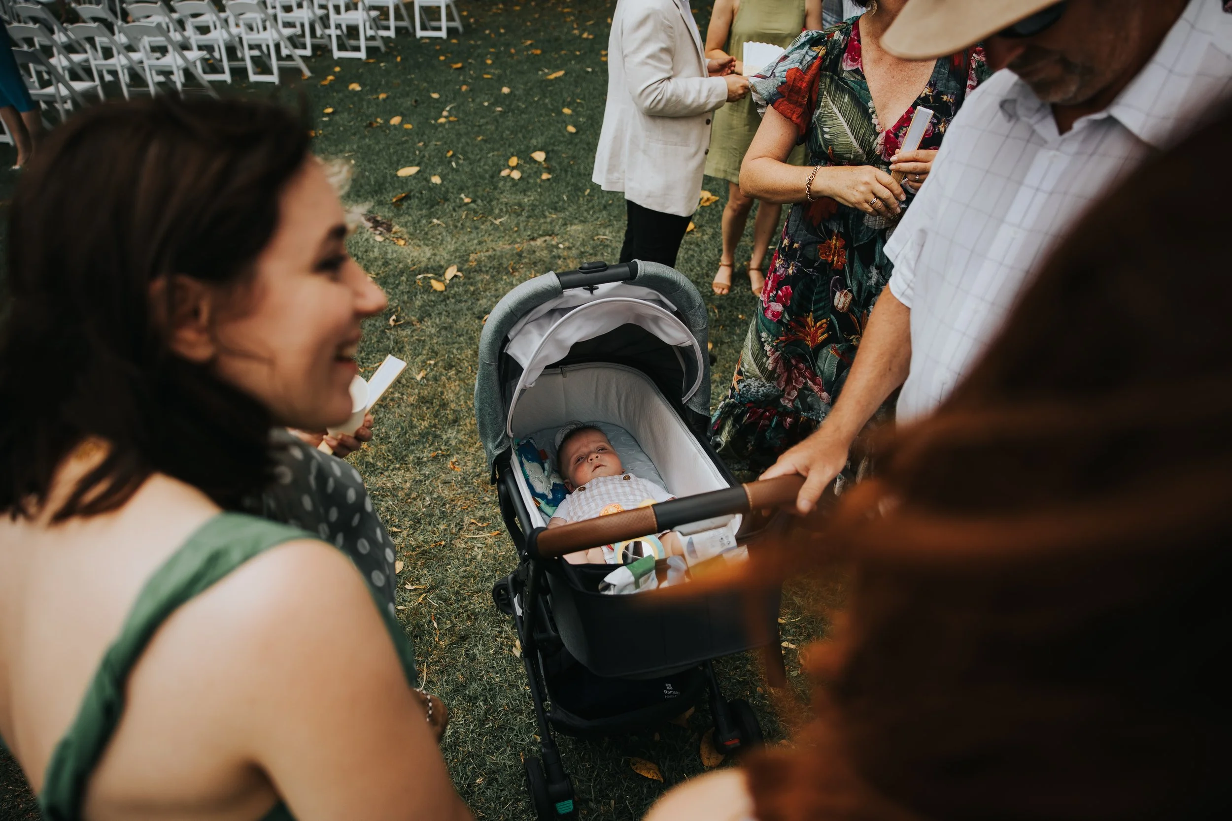 A group of people gathered outdoors at an event, with chairs set up and a baby in a stroller in the center. The baby is looking up, surrounded by adults engaged in conversation.