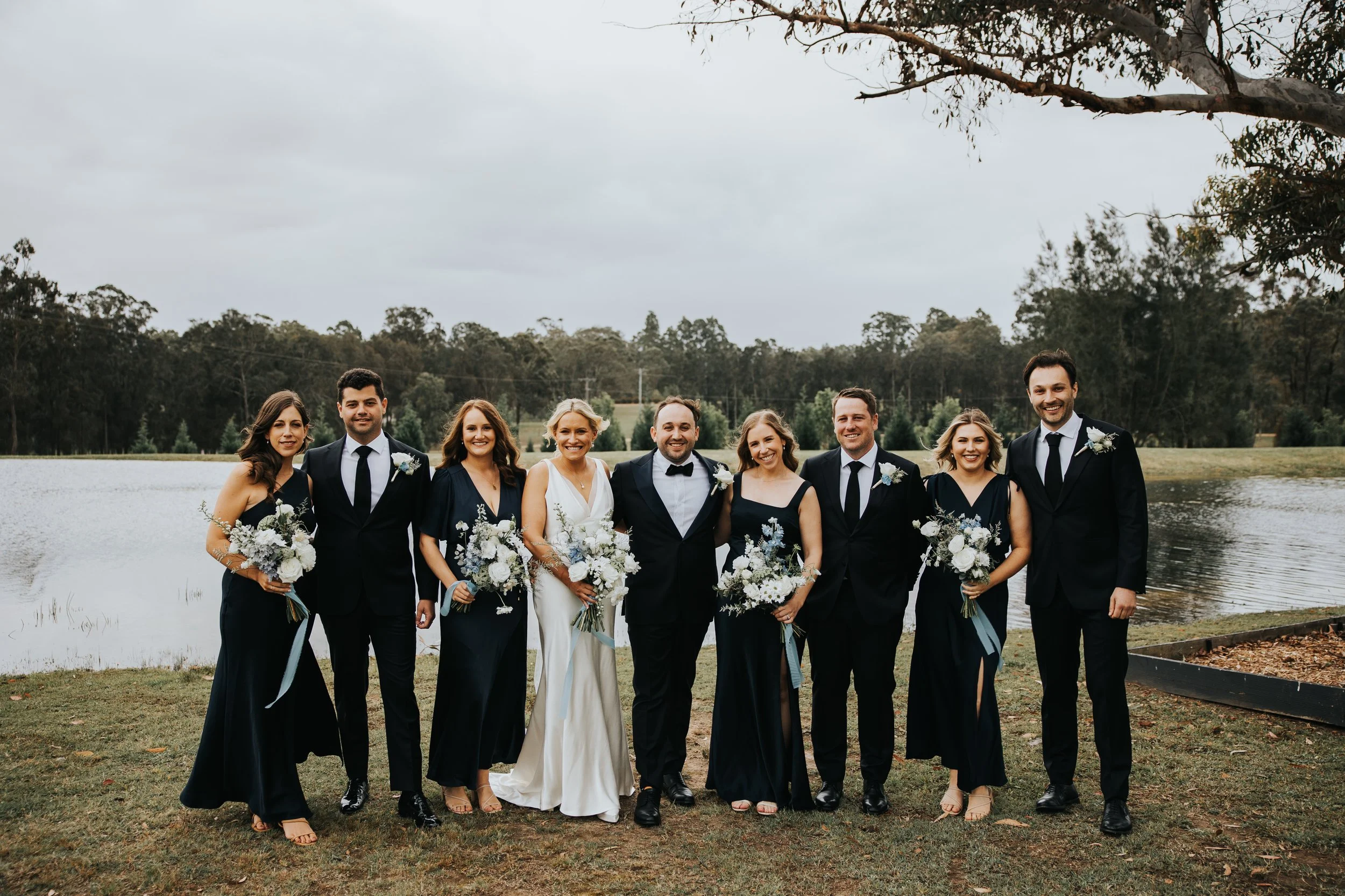 Group of nine people dressed in formal attire, including wedding dresses and suits, standing outdoors near a lake with trees in the background, celebrating a wedding.