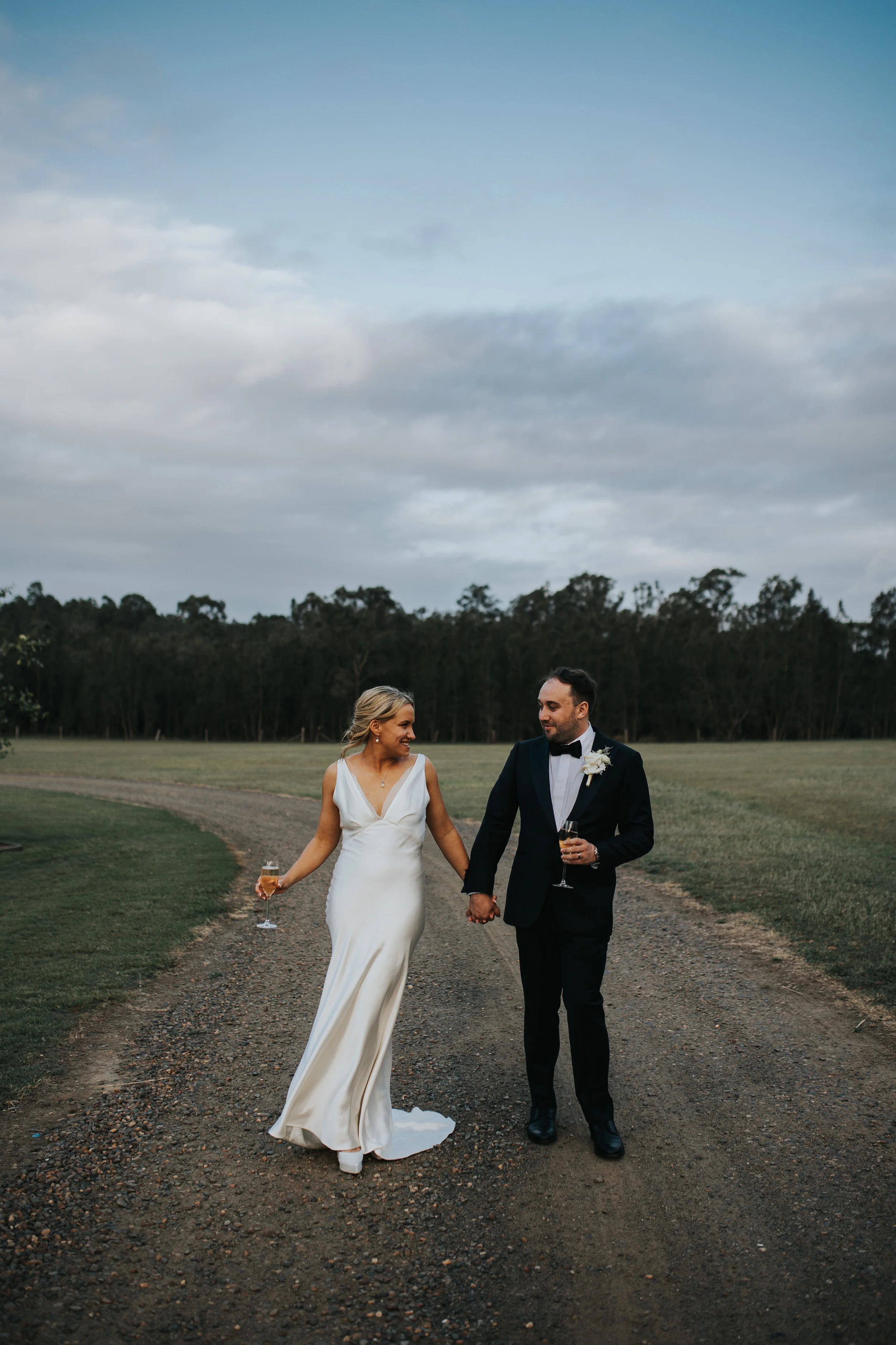 A bride and groom walking hand in hand on a gravel path outdoors during their wedding celebration, holding glasses of champagne, with trees in the background and cloudy sky overhead.