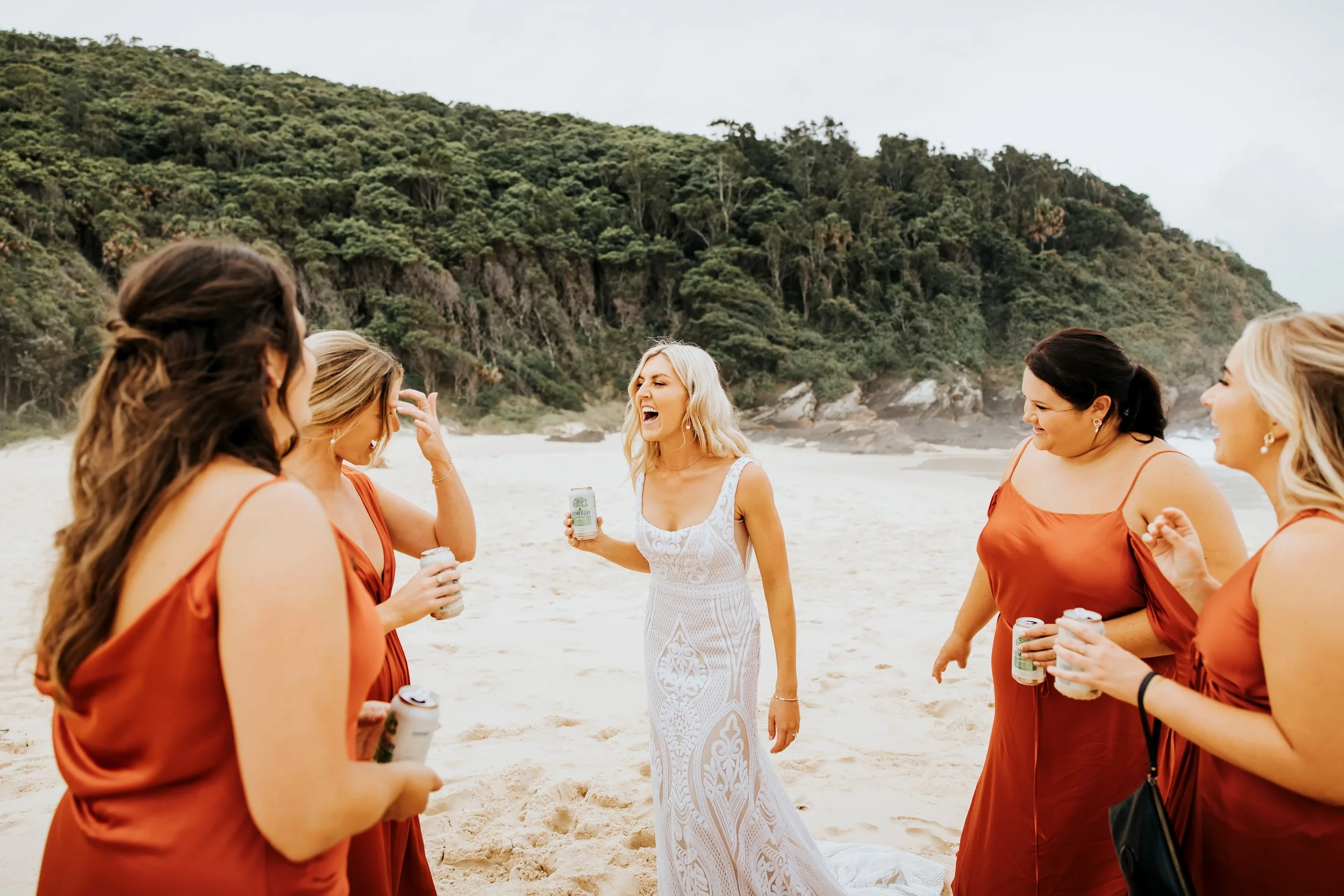 A group of women on a beach, laughing and holding drinks, with one woman in a white dress and others in orange dresses, and a green hillside in the background.