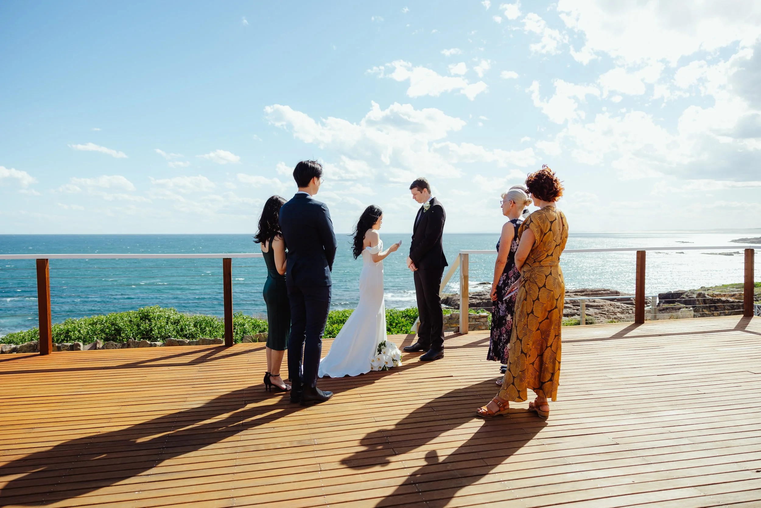 A wedding ceremony taking place on a wooden deck overlooking the ocean with cloudy blue sky. The bride and groom face each other while five guests, three women and two men, stand observing them. The bride wears a white dress and the groom wears a bla