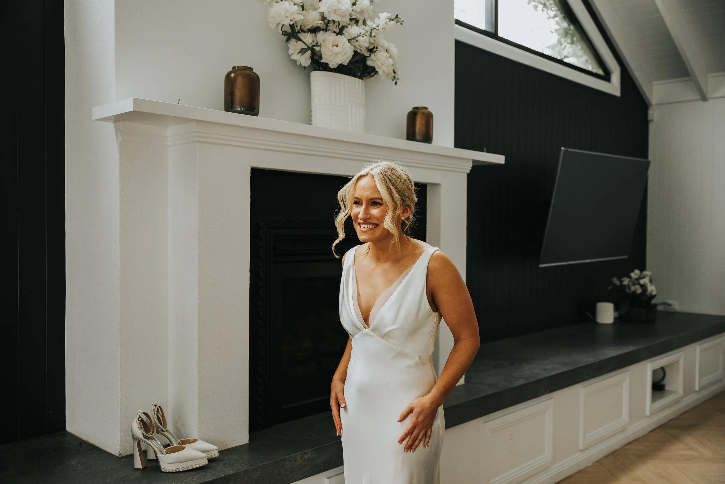 A woman in a white dress smiling in front of a fireplace with a large white flower bouquet on the mantel and two brown vases. Shoes are next to the fireplace on the floor.