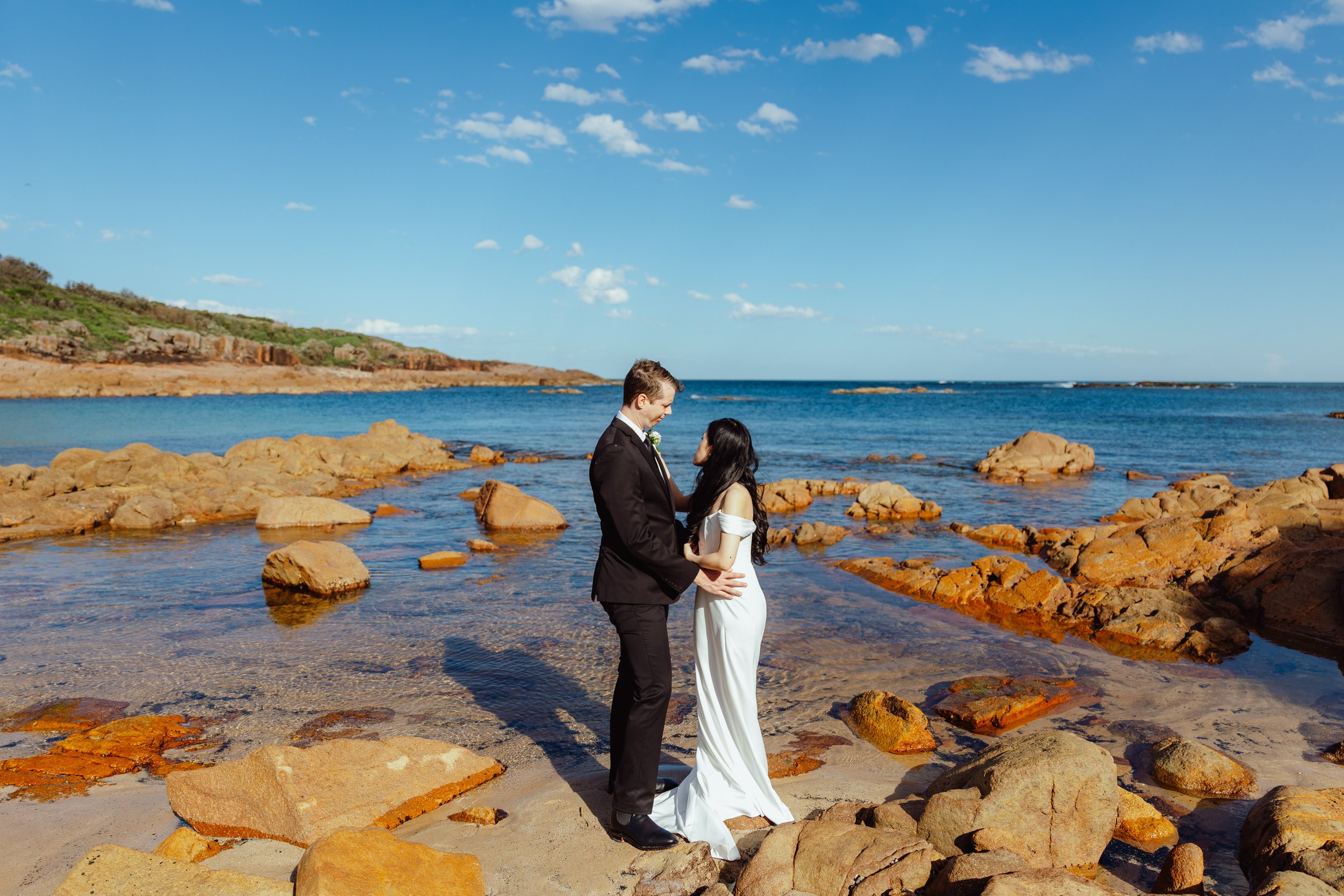 A couple in wedding attire standing on rocky beach by the ocean with blue sky and scattered clouds.