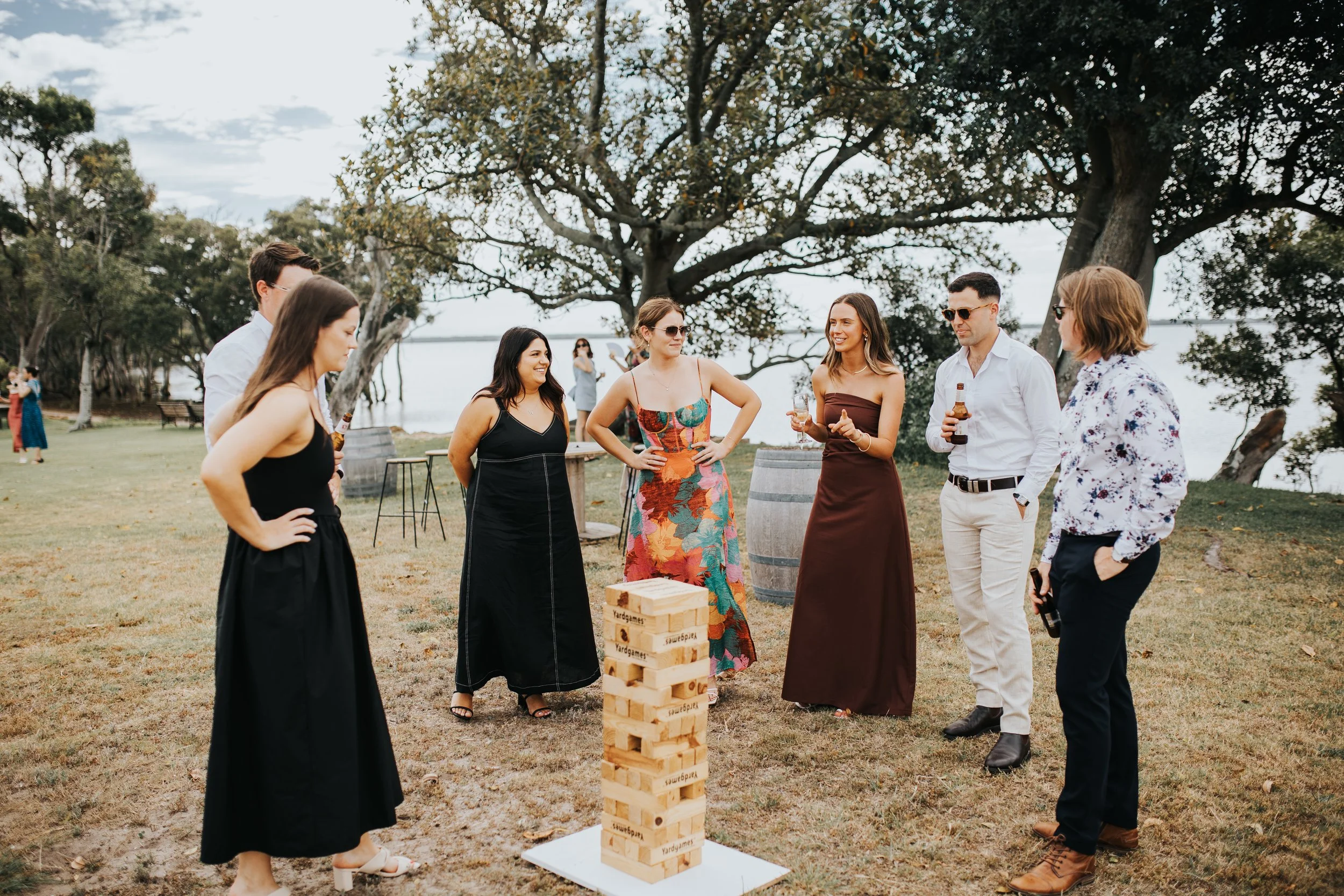 Group of people gathered outdoors playing giant Jenga during daytime with trees and water in the background.