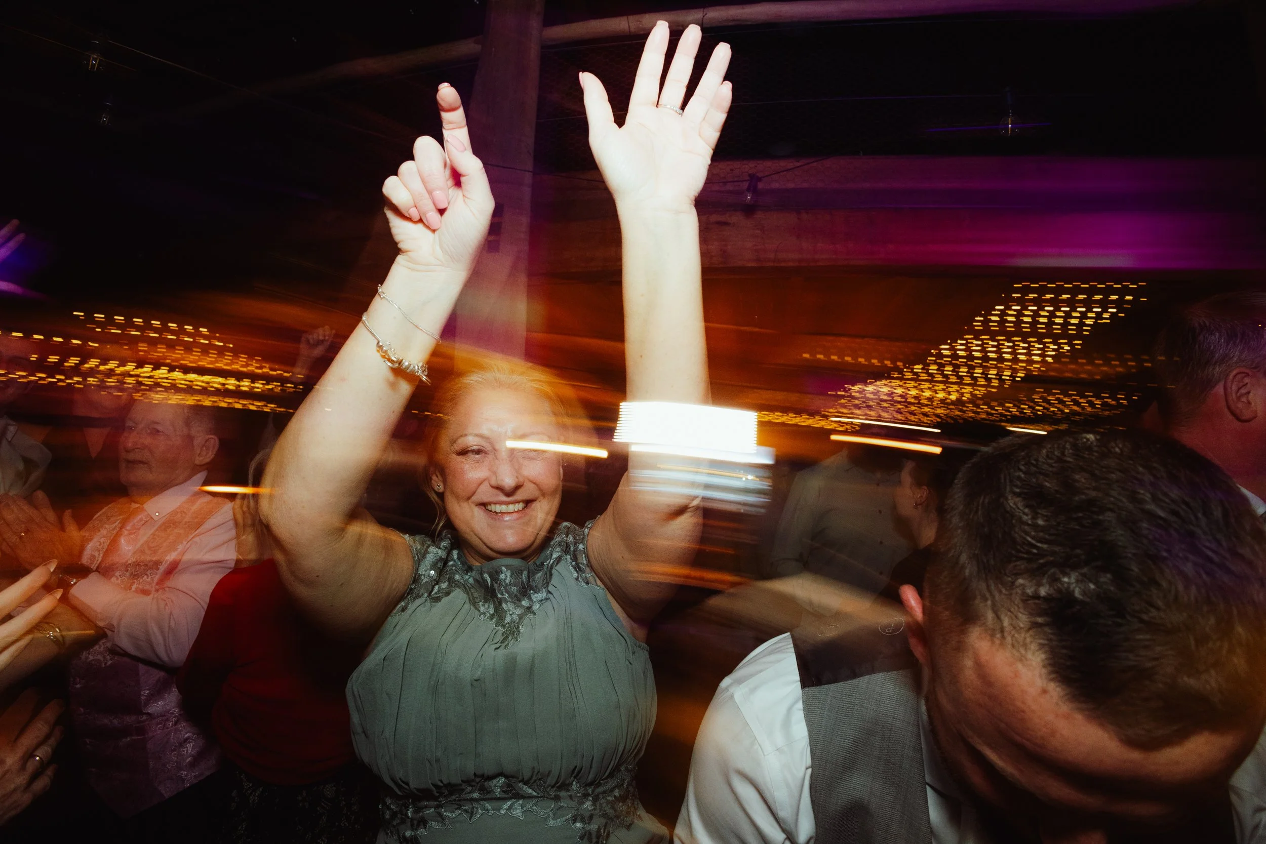 A woman smiling and dancing at a lively party or celebration, raising her hand with a peace sign. Other people are visible in the background, and colorful lights create a festive atmosphere.