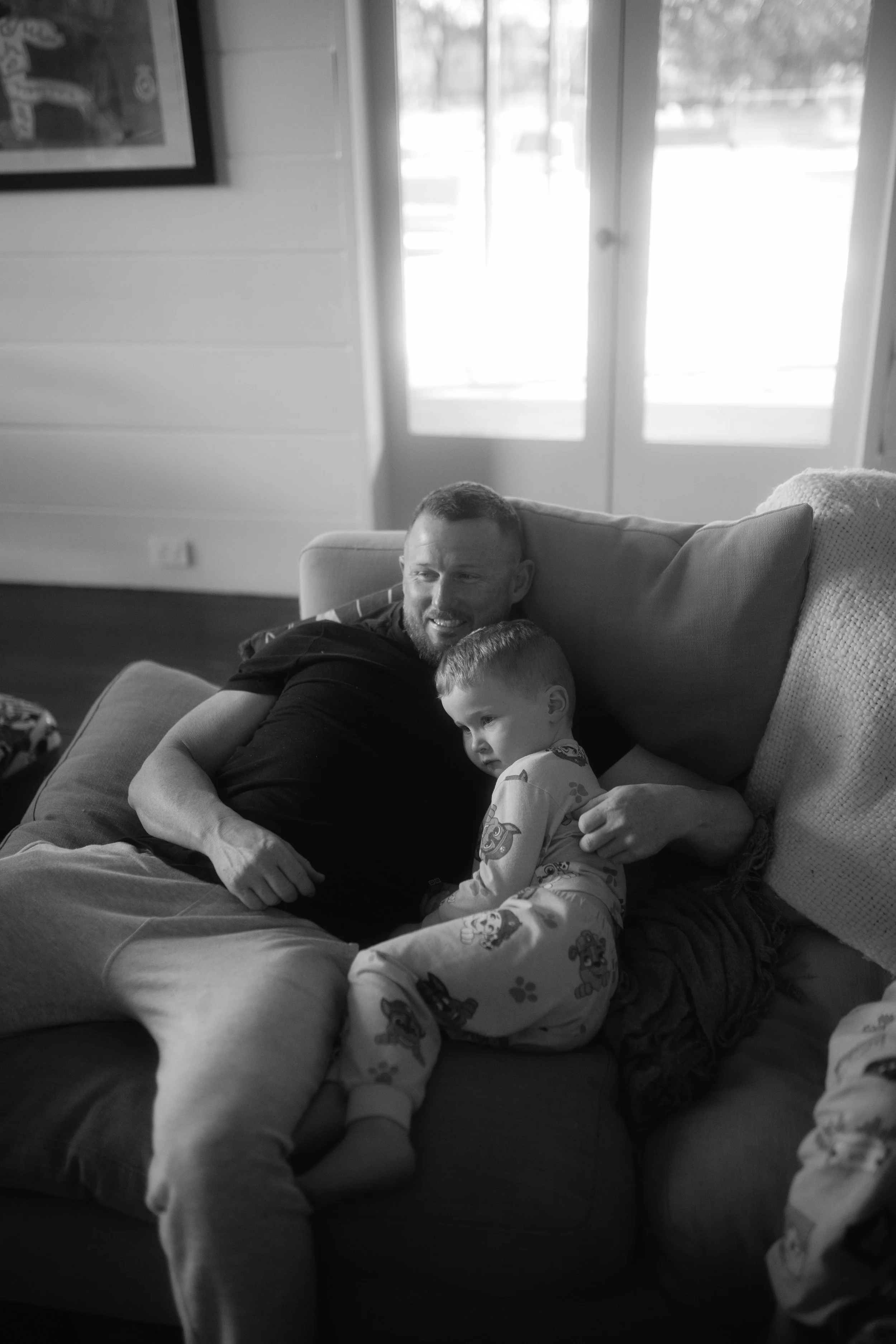 A man with a beard and short hair sitting on a couch with a young boy who has his head resting on the man's chest. They are inside a room with large windows in the background, allowing natural light to fill the space.