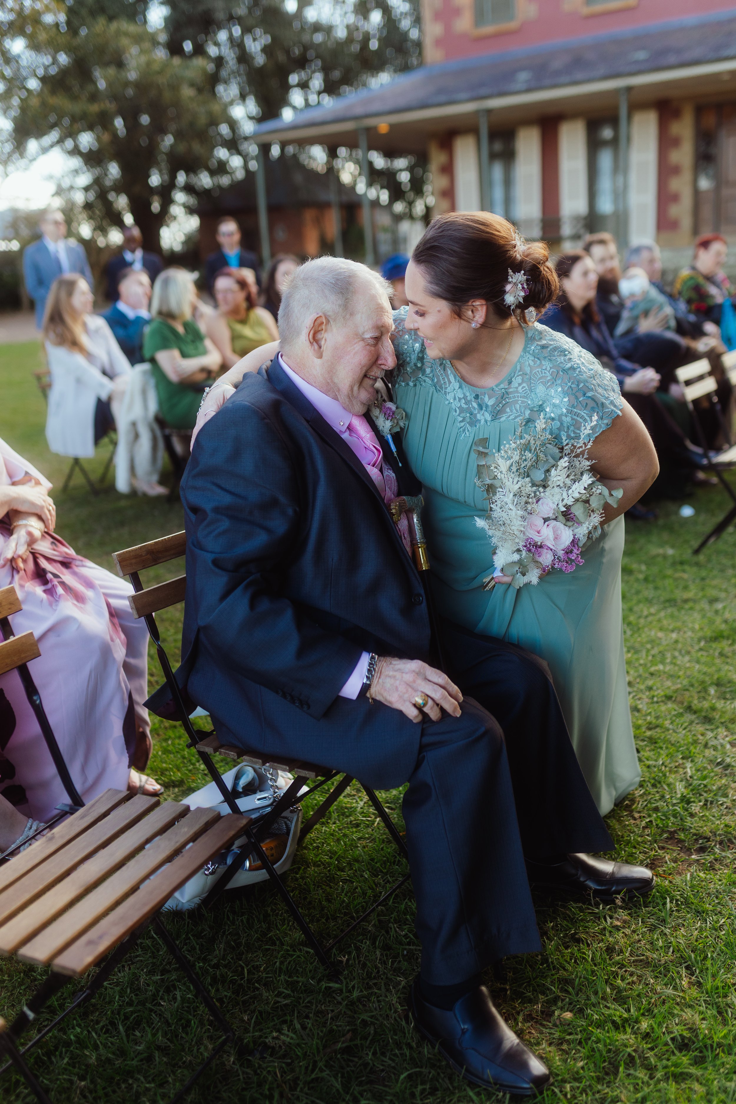 A woman in a blue dress leaning in to kiss an elderly man in a suit, seated outdoors at a wedding ceremony, with guests seated in the background.