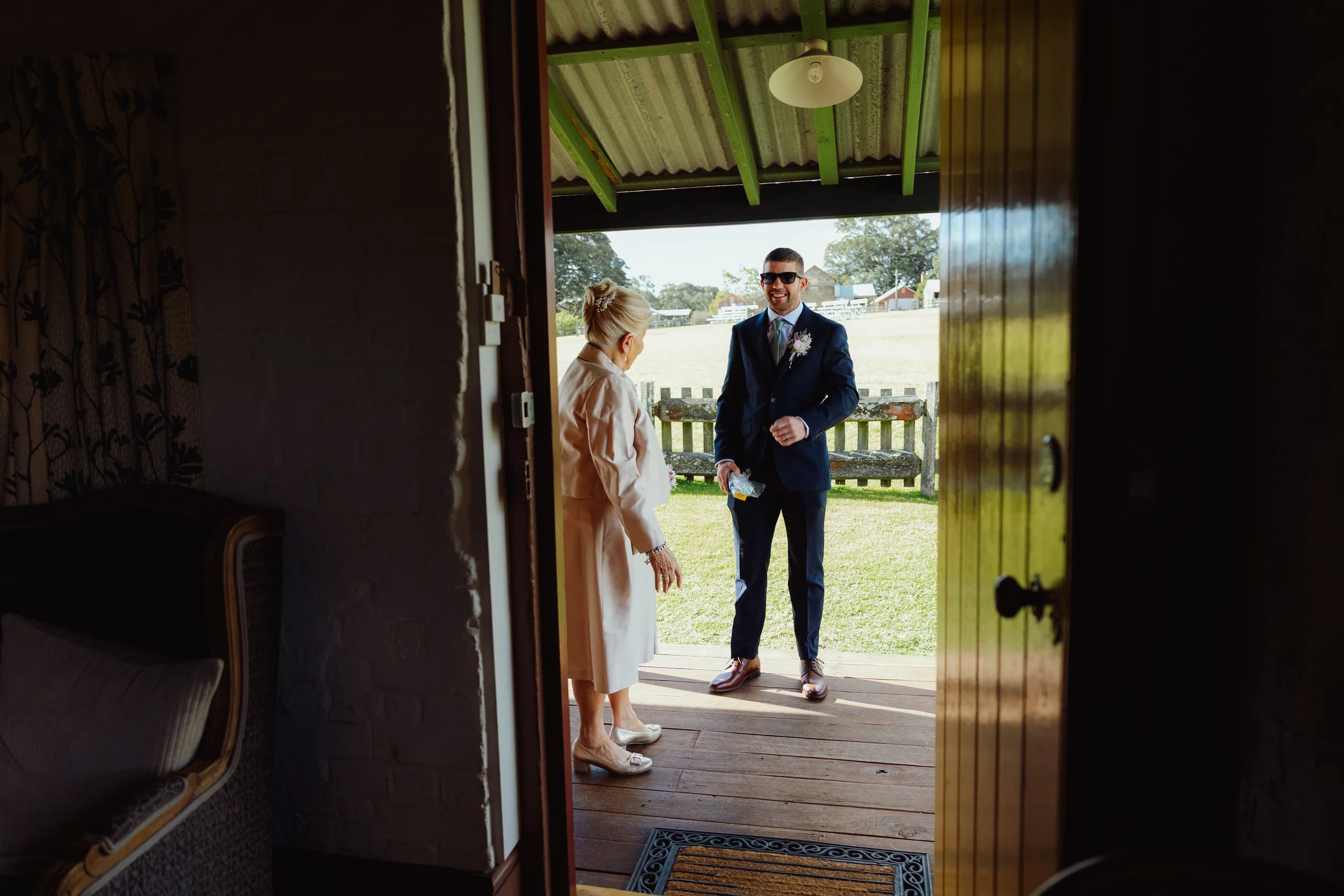 A man in a navy suit, sunglasses, and a boutonnière greets a woman in a beige dress and heels at the door of a rustic outdoor setting, with a field and farm buildings in the background.