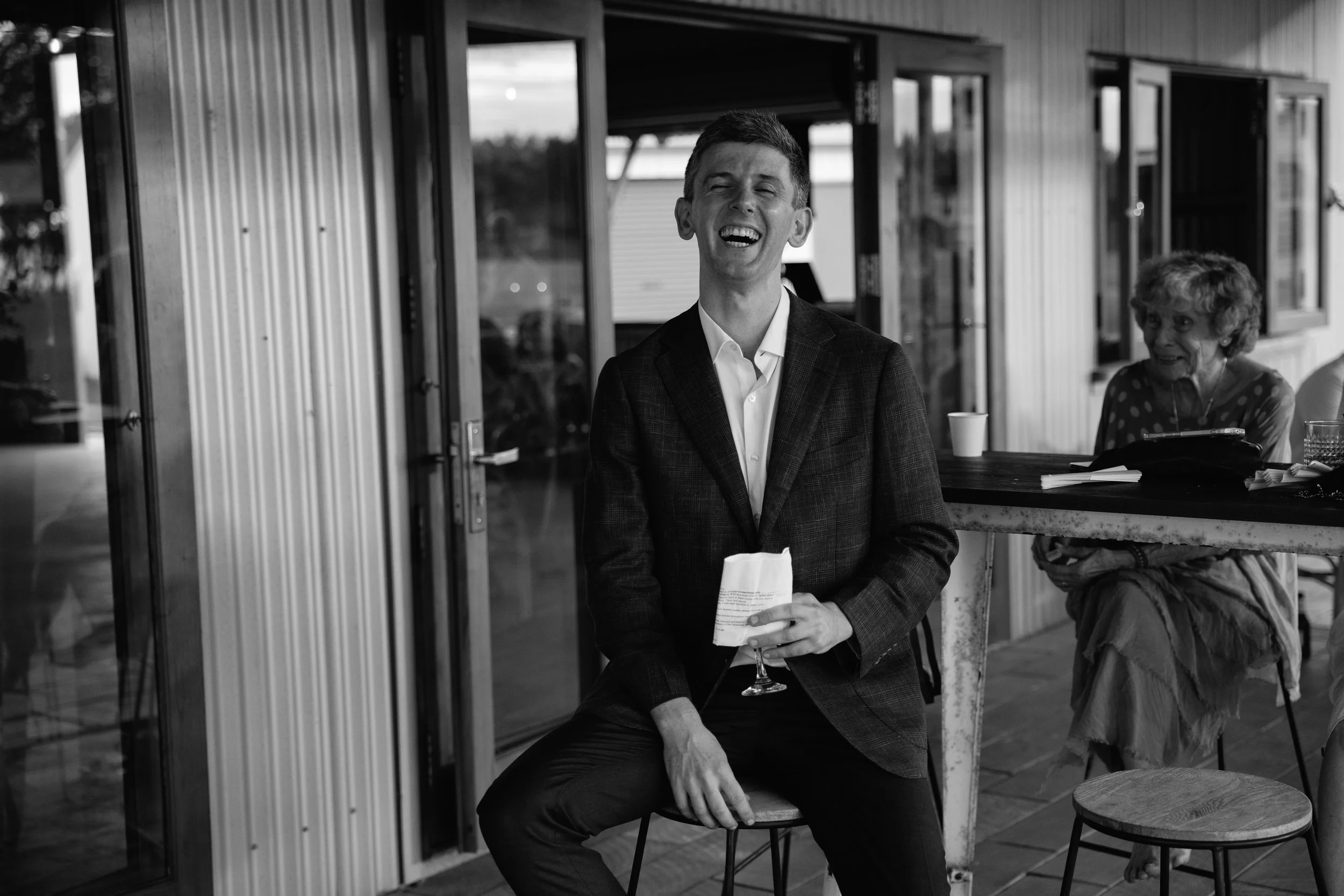 A man dressed in a suit sitting at a table outdoors, laughing with his eyes closed, a woman with curly hair and a polka-dot shirt sitting nearby, and a window in the background.