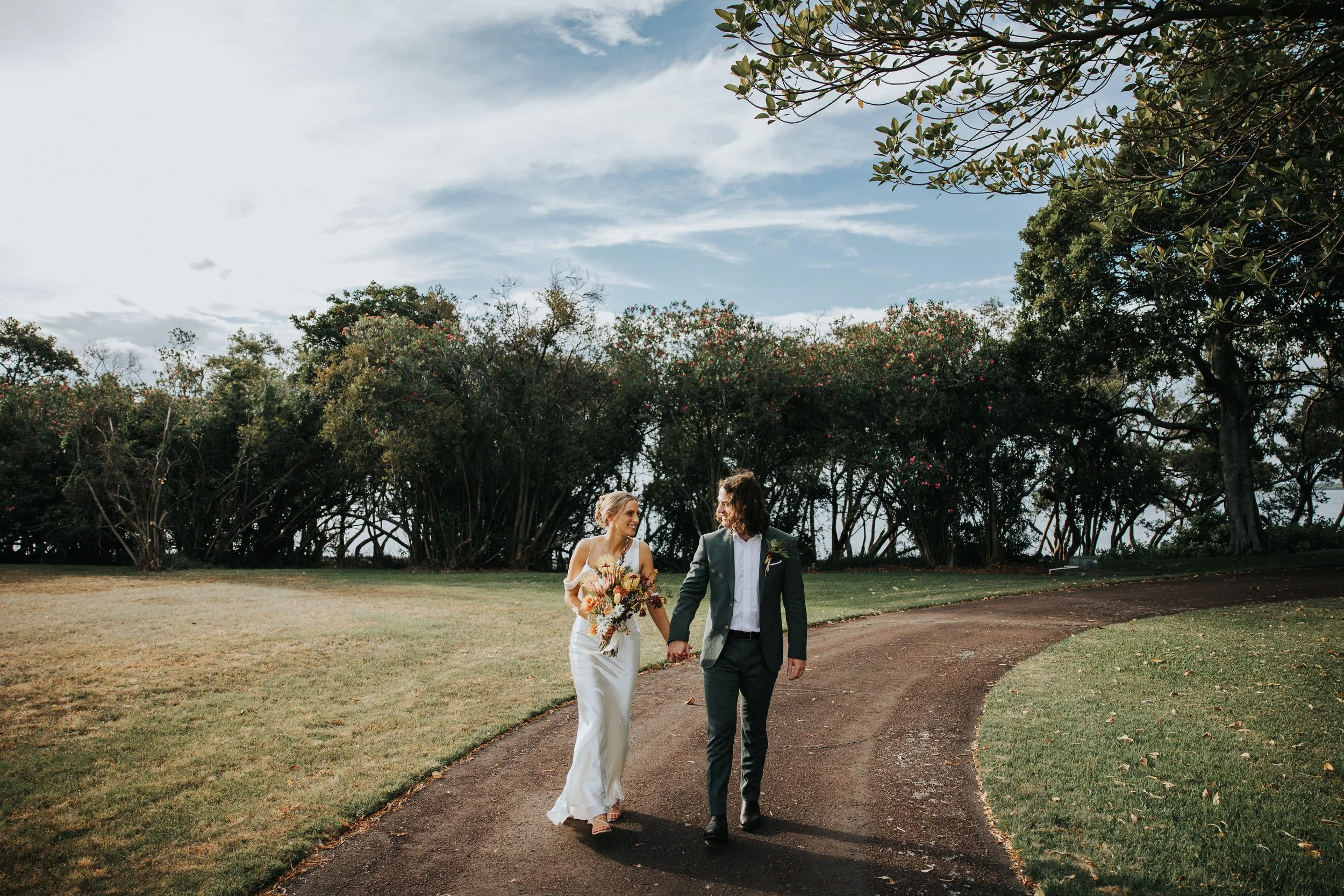 A bride and groom holding hands and walking outdoors on a curved path, surrounded by trees, during their wedding day.