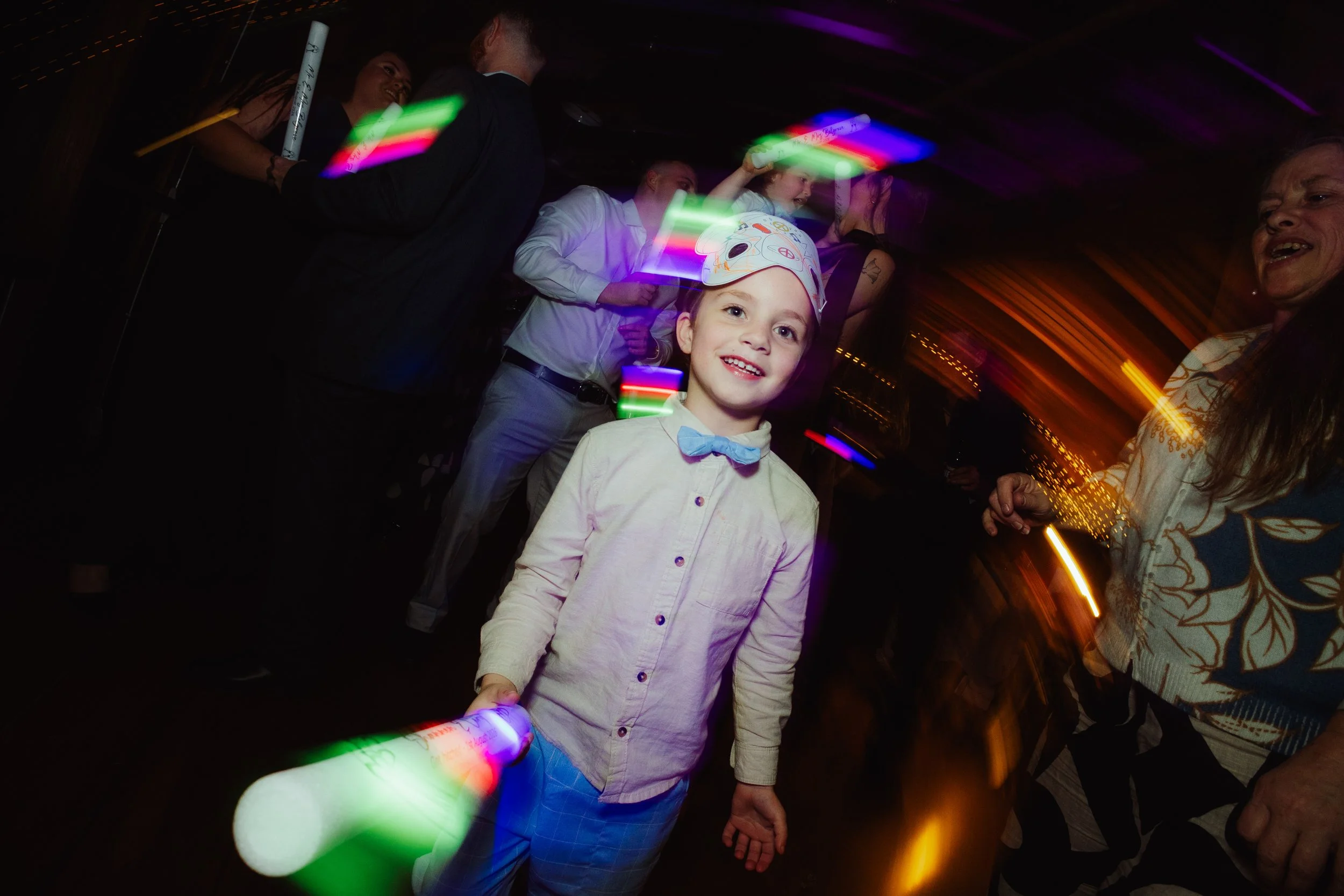 Young girl wearing a party hat and bow tie holding a glow stick at a party with people dancing around in a dark room illuminated by colorful lights.