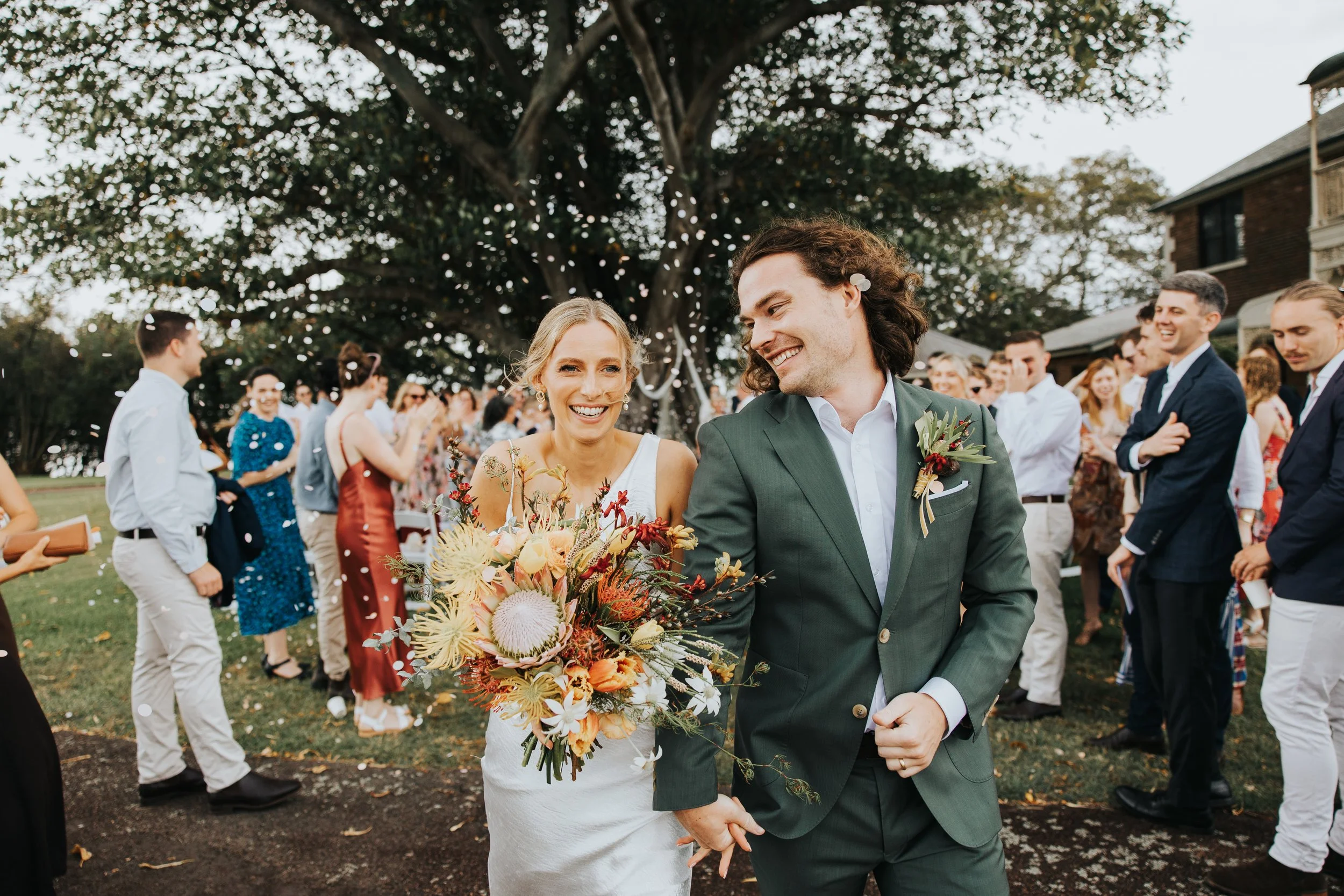 A bride and groom holding hands and smiling during their outdoor wedding celebration under a large tree, surrounded by guests. The bride is holding a large bouquet of colorful flowers, and the groom is wearing a green suit with a boutonniere, both ap